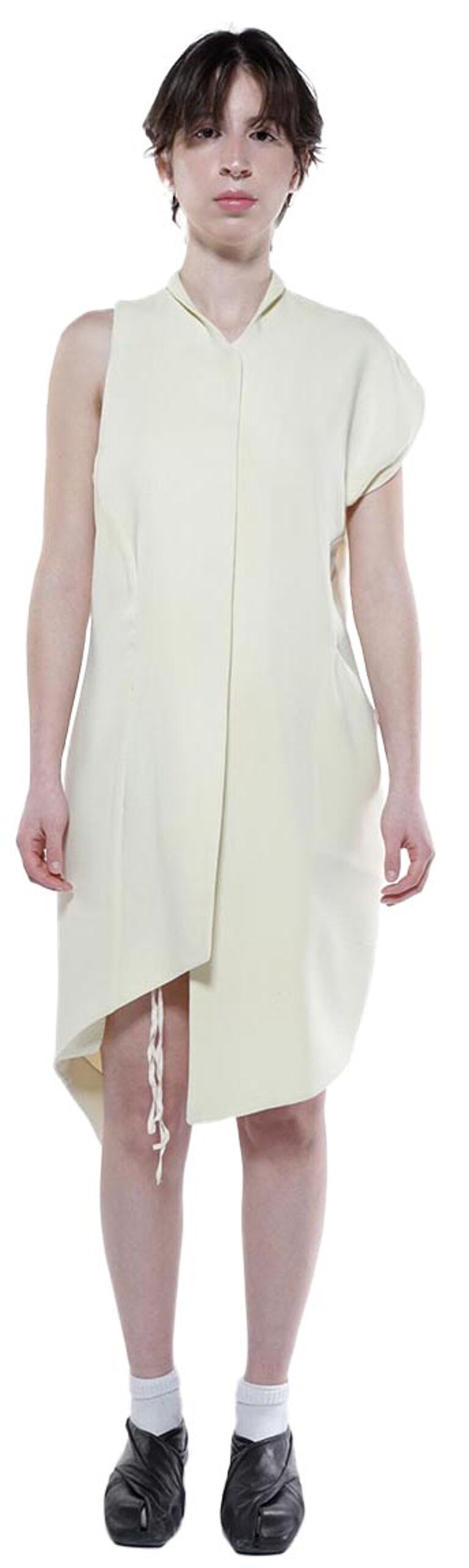 A person with short brown hair wears a pale yellow asymmetric sleeveless dress and dark shoes.