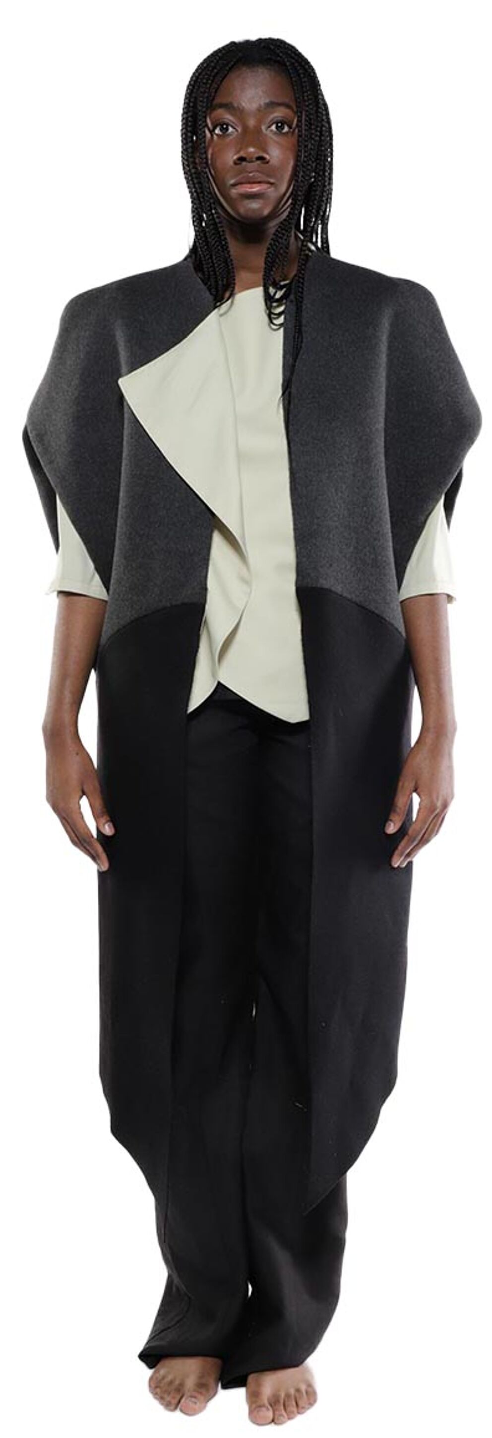 A person with dark skin and long black braids wears a gray and black asymmetrical long vest over a light beige top and black pants.