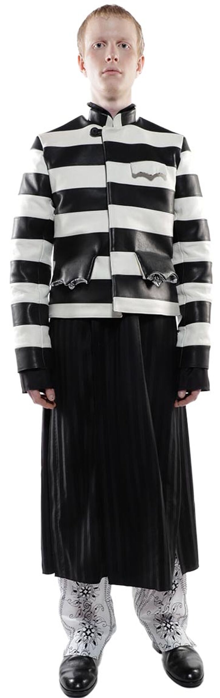 A person with fair skin and short hair wears a black and white striped jacket, a long black skirt, and patterned boots.