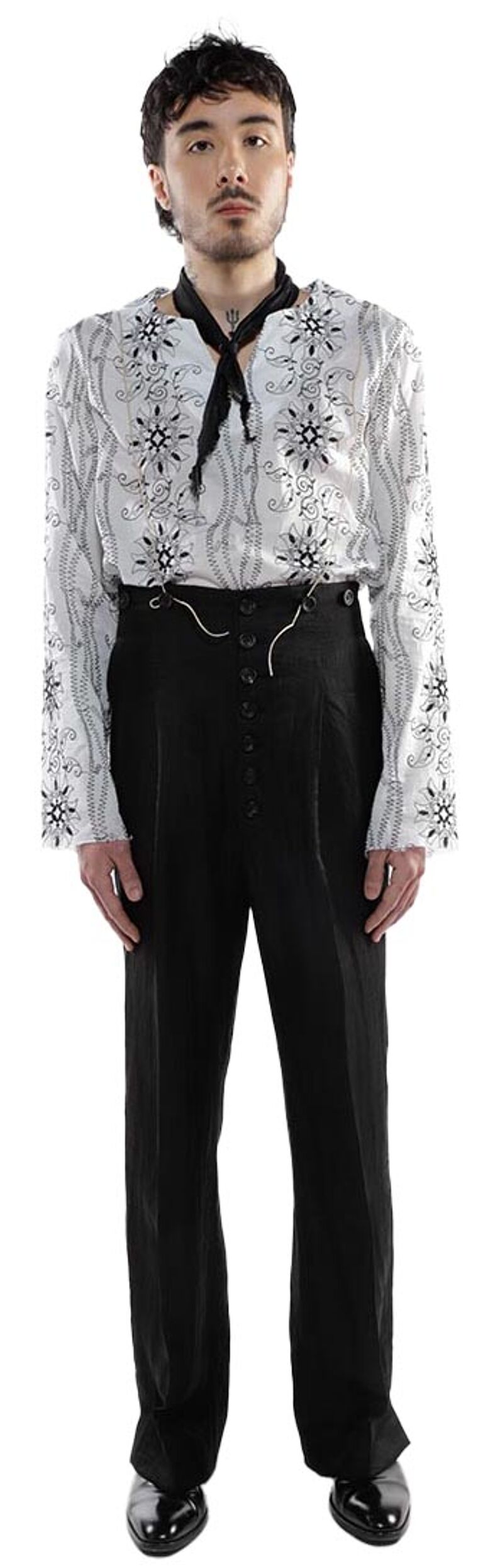 A person with dark hair, wearing a white, patterned long-sleeved shirt, black pants, and black shoes, stands against a white background.