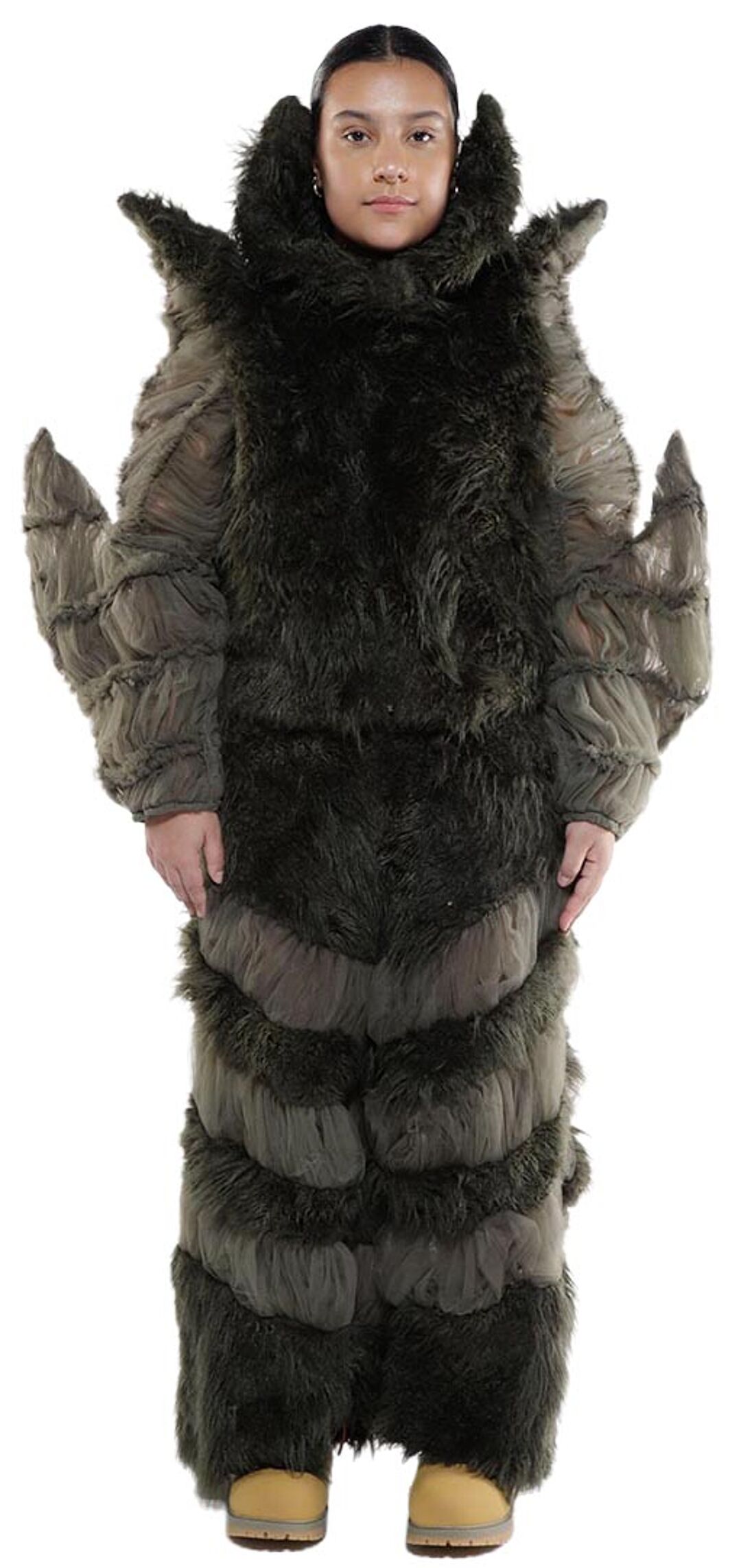 A person wearing a dark gray furry costume resembling a segmented creature stands against a white background, wearing tan boots.