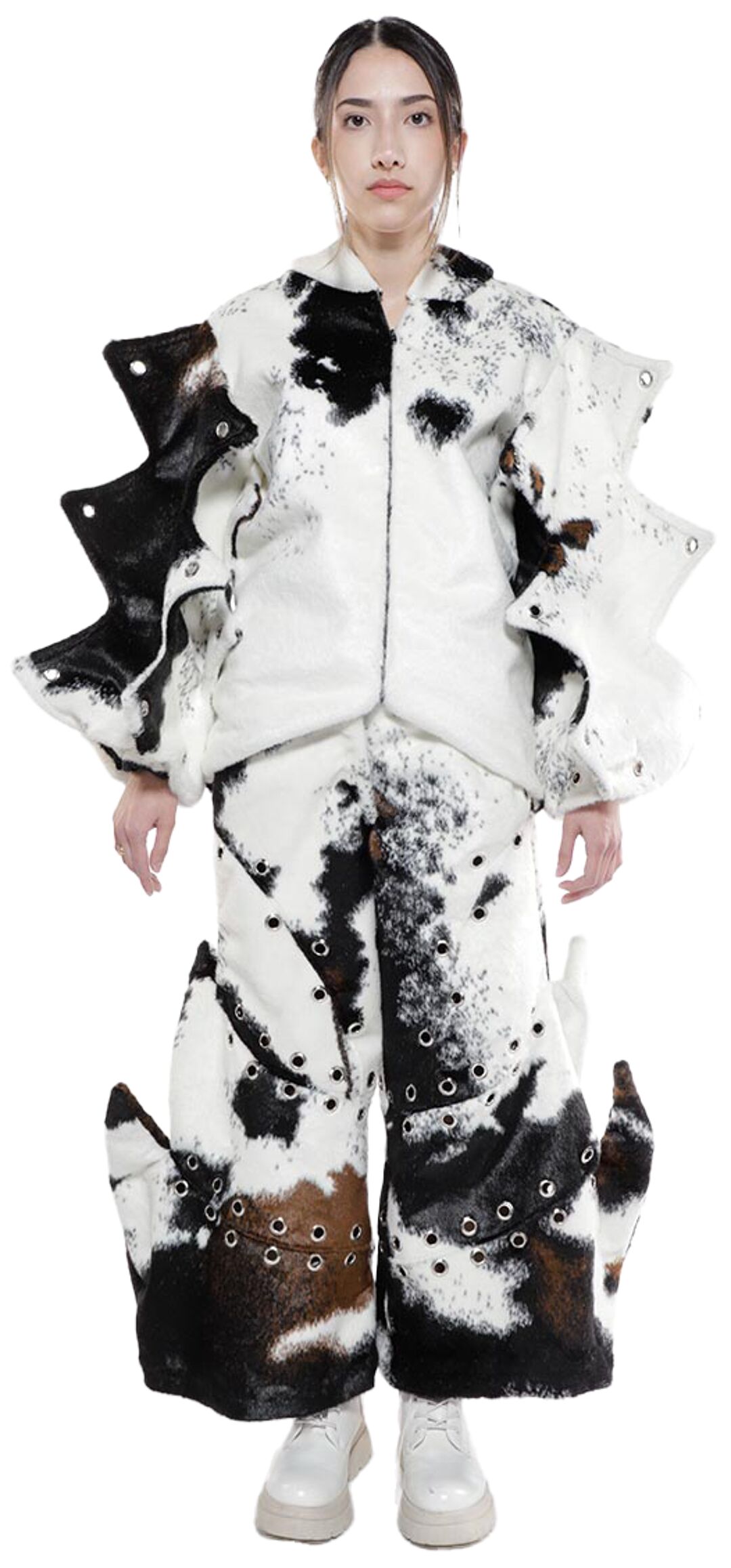 A young woman with dark hair pulled back models a unique, cow-patterned, avant-garde outfit consisting of a jacket and wide-legged pants with exaggerated detailing and grommets, paired with white lace-up boots, set against a white background.