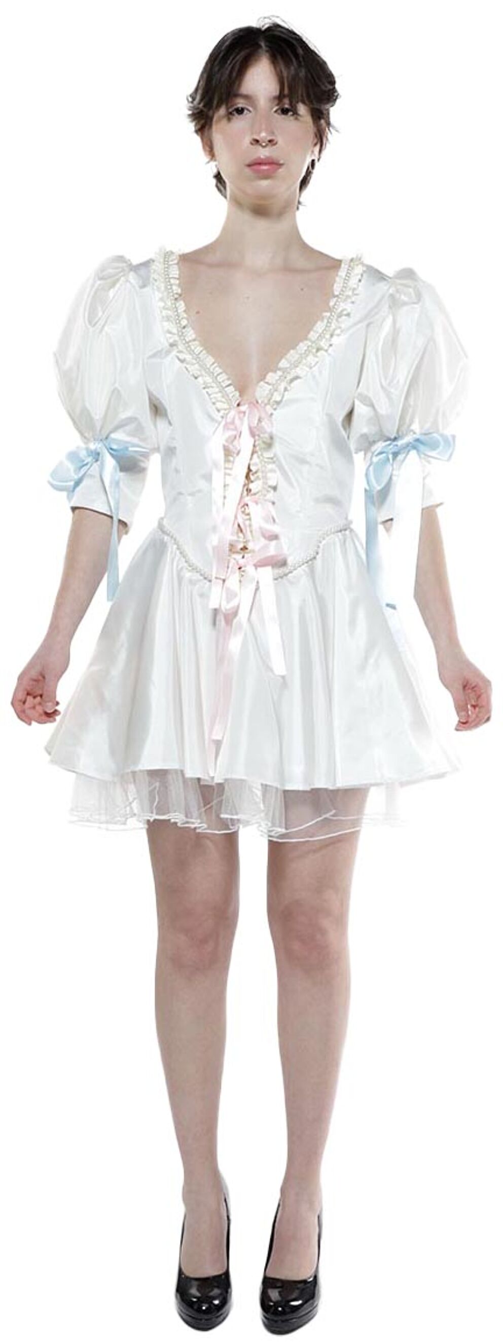 A young woman with dark hair, wearing a white satin dress with light blue and pink ribbons, and black heels, stands against a white background.
