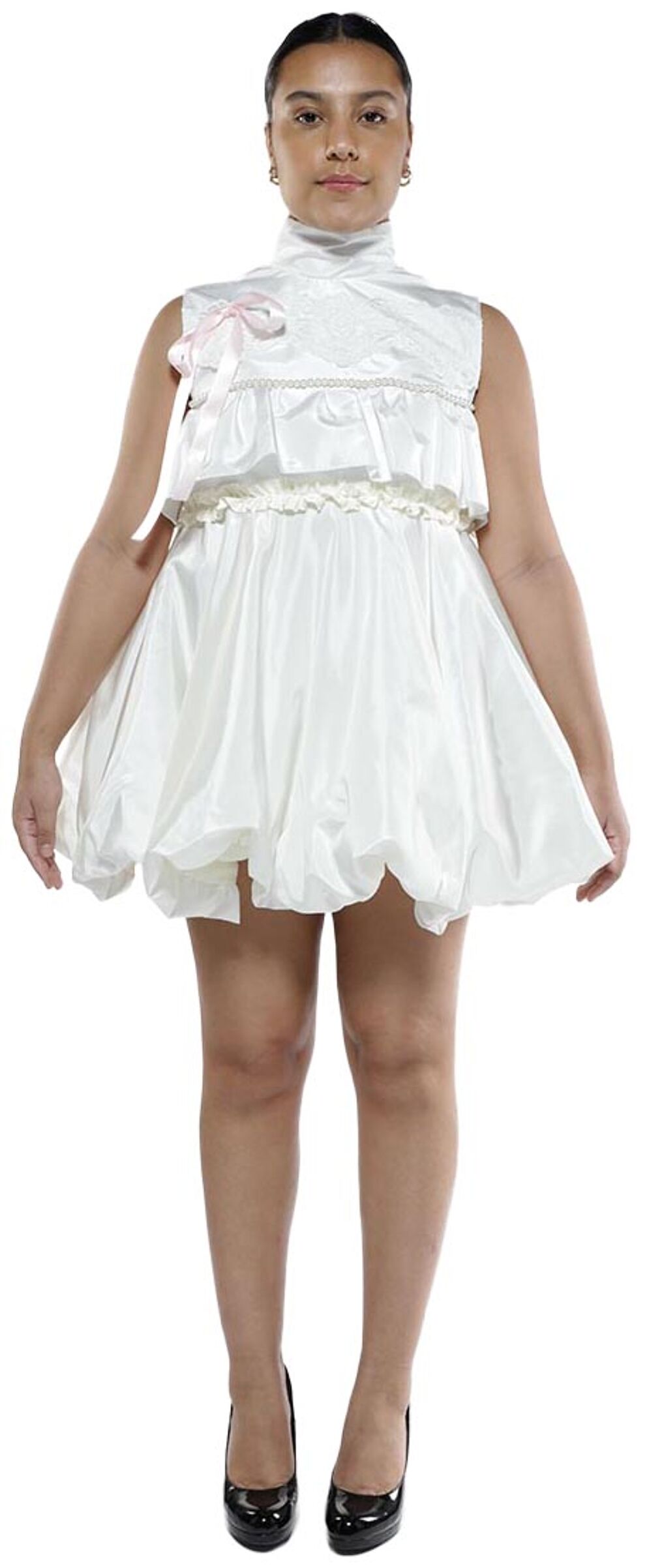 A young woman with tan skin and dark hair is standing and wearing a white, puffy, satin mini-dress with a high neck and black heels.