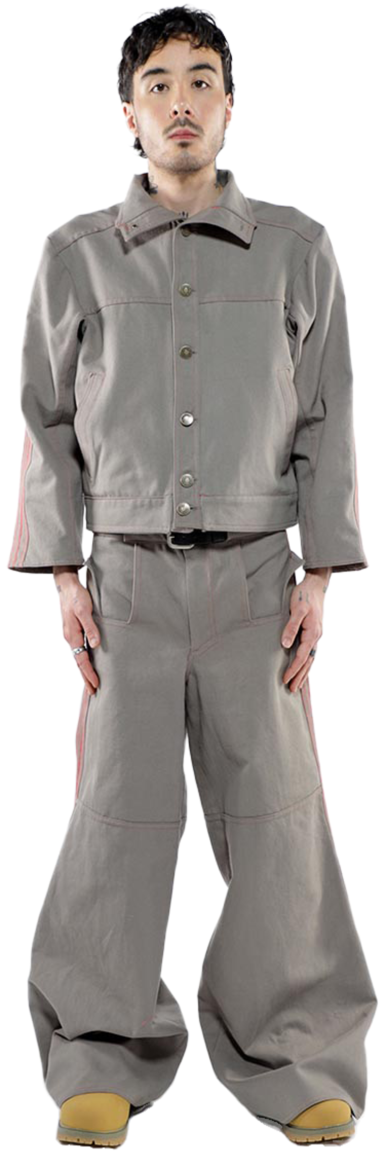 A young man stands against a white background wearing a taupe button-up jacket and matching wide-leg pants with thin pink stripes down the sides.