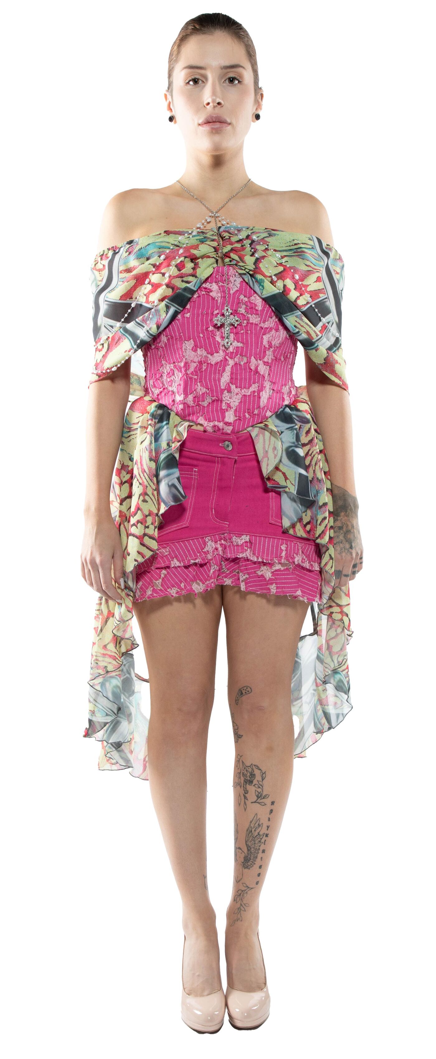A woman with short, light brown hair and tattoos on her legs wears an off-the-shoulder multicolored top over a pink corset and skirt, paired with nude heels.