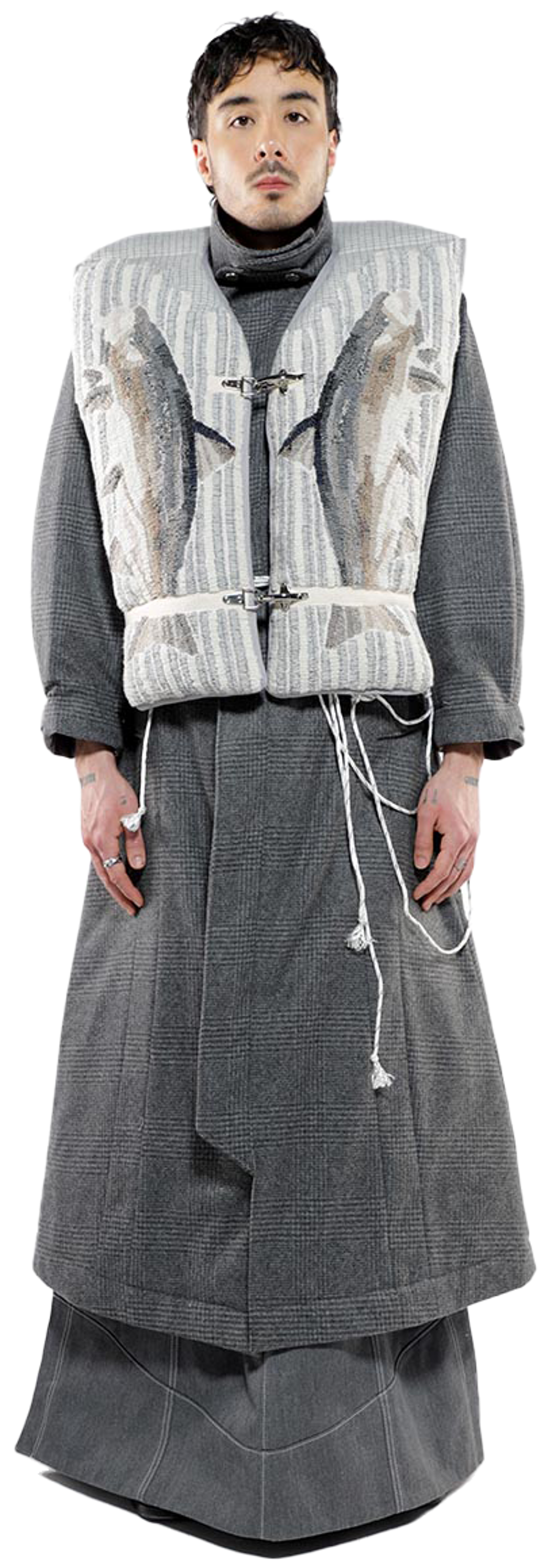 A person with dark hair and a neutral expression wears a long gray coat over a lighter gray vest with embroidered fish, standing against a white background.