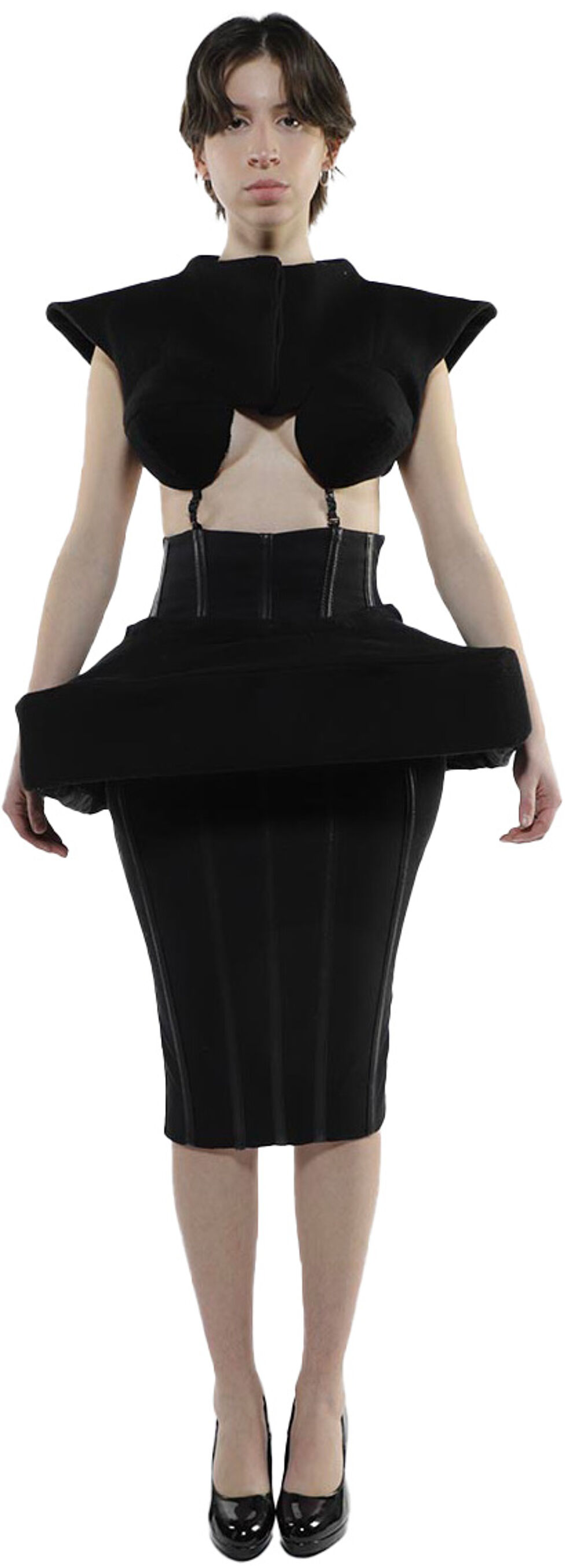 A woman with short dark hair is standing and wearing a black, form-fitting outfit with a unique, structured bodice and skirt, and black high-heeled shoes.