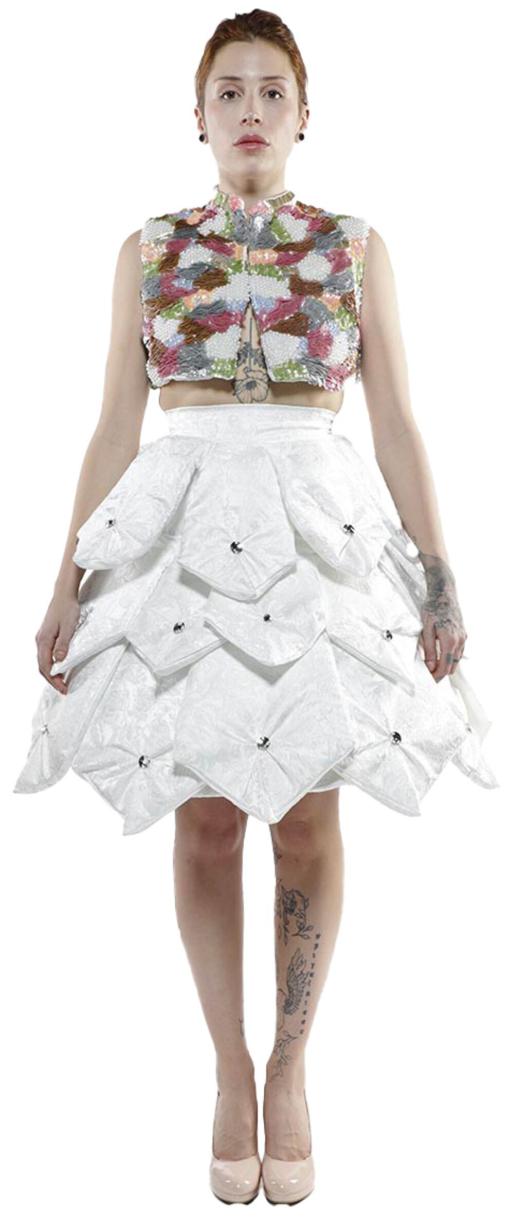 A woman with reddish-blonde hair and tattoos on her legs is posing in a white sequined top and a layered white skirt adorned with rhinestones, wearing nude heels.