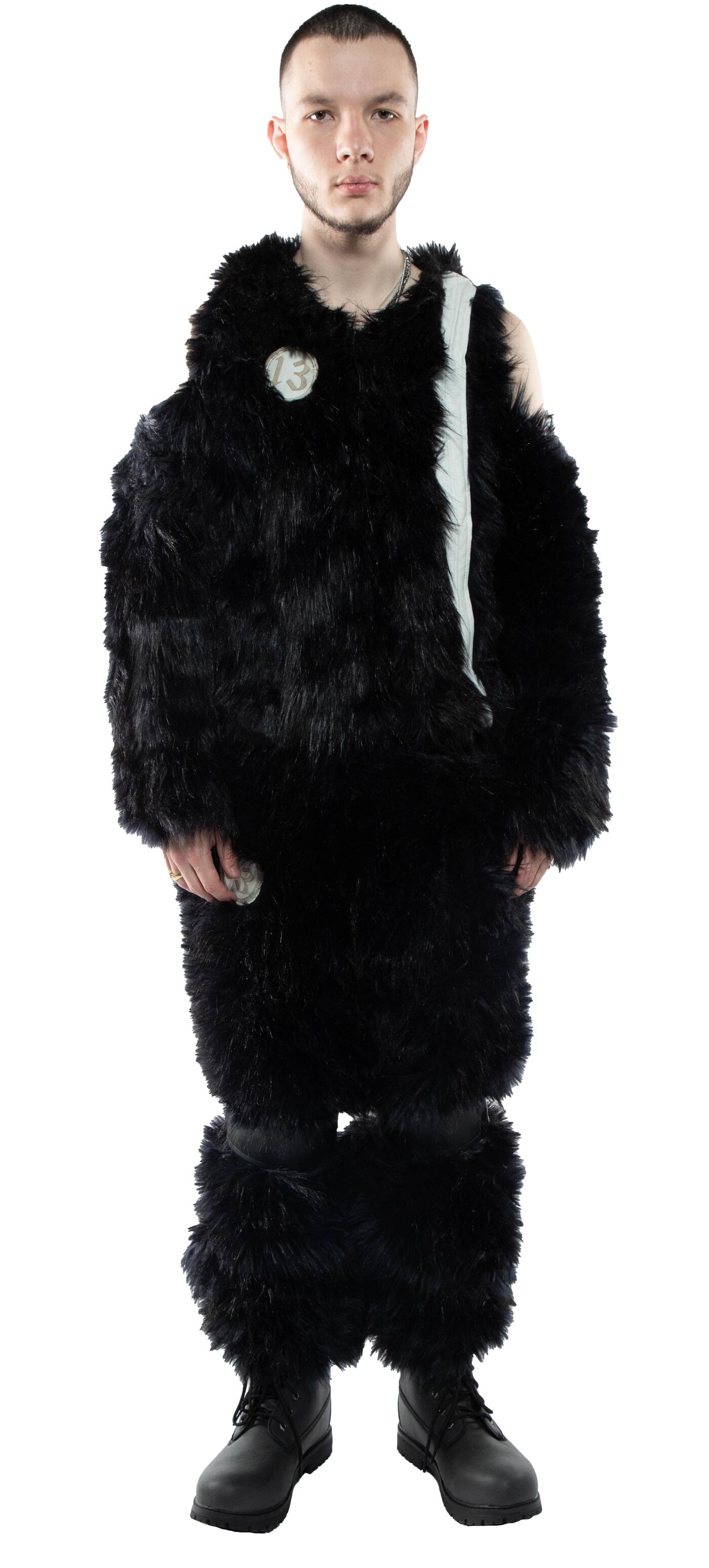 A person with short brown hair is modeling a black fuzzy outfit consisting of a long coat, pants, and leg warmers, paired with black boots.