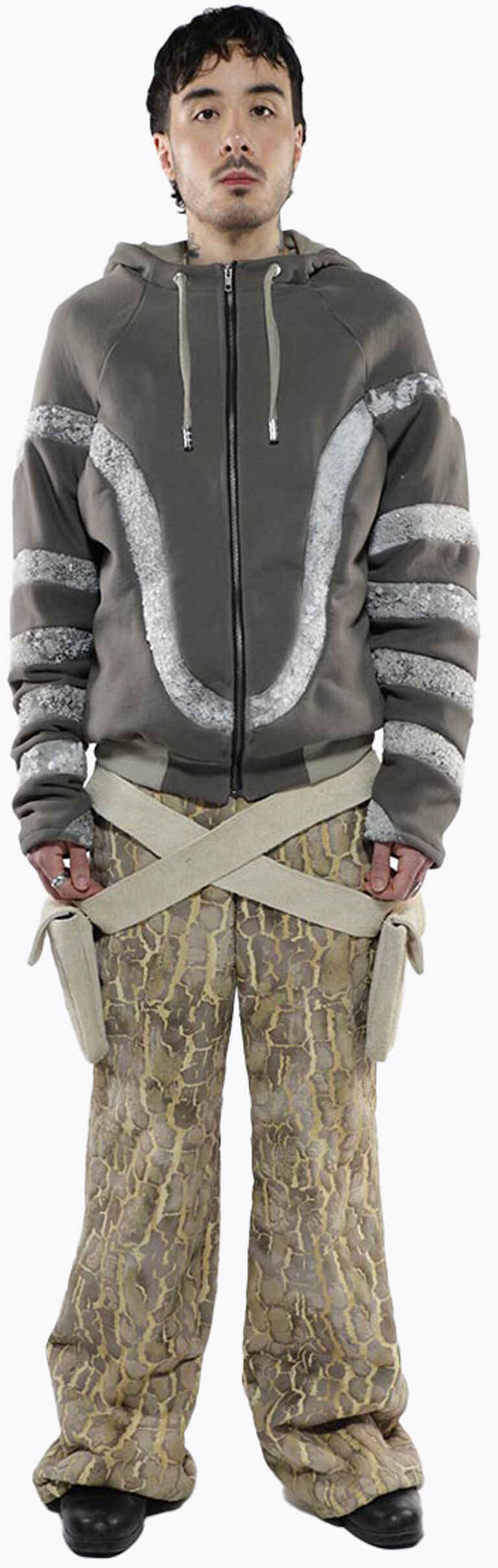 A man is standing, wearing a gray zip-up hoodie with white accents, beige pants with a pattern, and a beige cross-body strap.