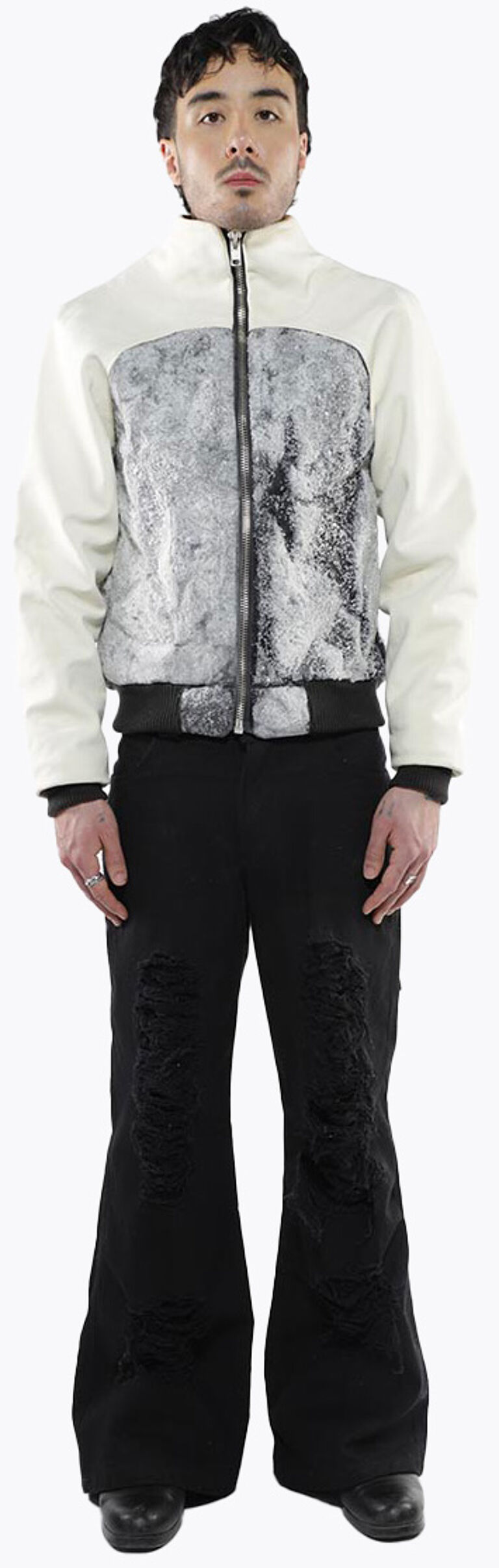 A young man with short brown hair stands against a white background wearing a white and gray bomber jacket and distressed black bell-bottom pants.