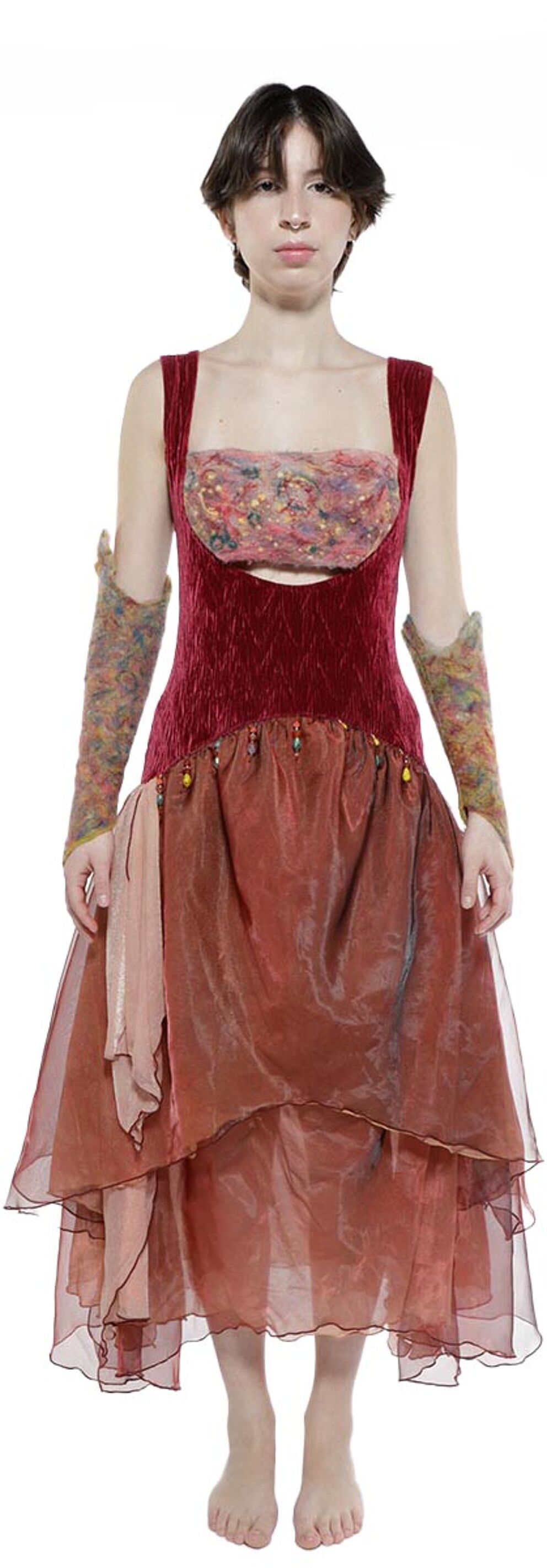A young woman with short brown hair wears a layered, rust-colored dress with long sleeves and sheer overlay, accessorized with textured arm warmers.