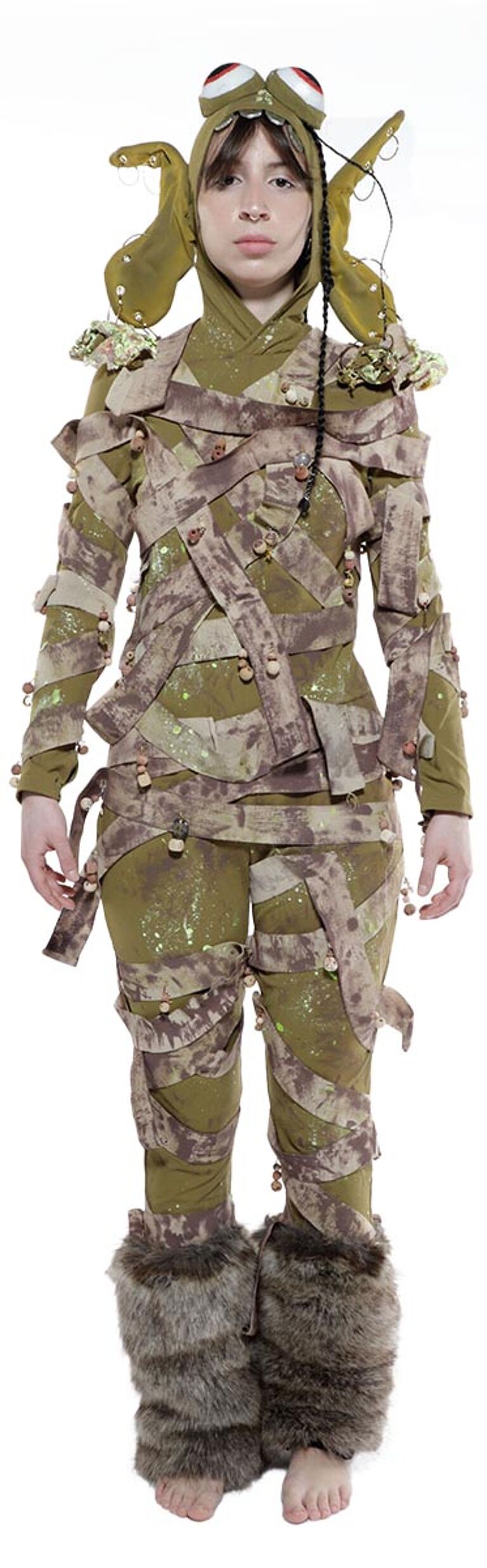 A child wearing a whimsical, tattered, olive-green costume with a frog-like hood, furry leg warmers, and numerous small bells is captured in a full shot against a stark white background.