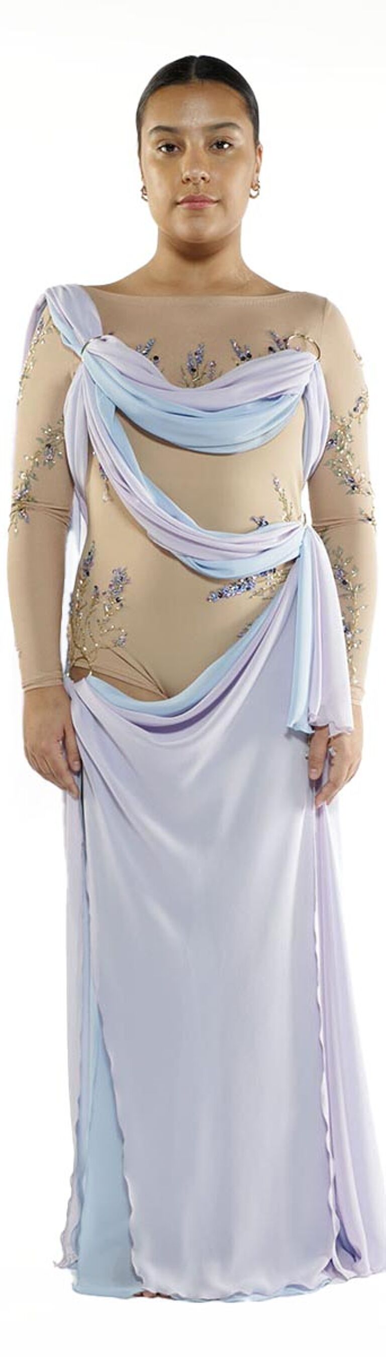 A woman with dark hair pulled back wears a long, draped gown in shades of beige, lavender, and light blue, with delicate embellishments.