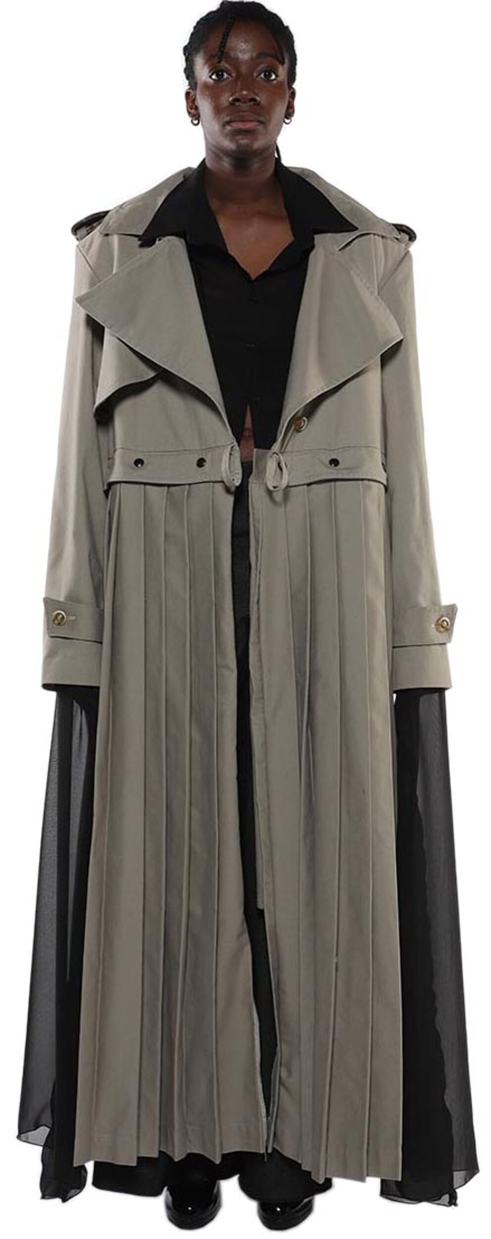 A model stands wearing a pleated, taupe-colored trench coat with black chiffon sleeves and a black collared shirt underneath.
