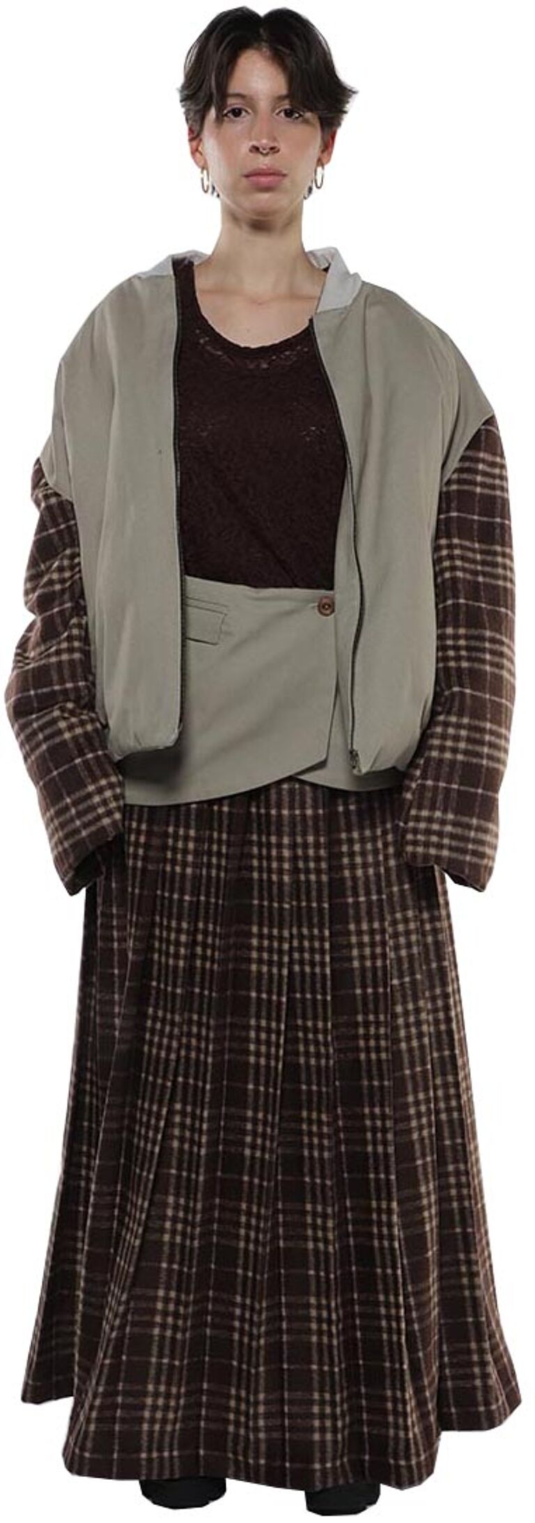 A person with short brown hair is wearing a brown and beige plaid pleated skirt and a matching jacket with a light green puffer vest over it, a brown top underneath.
