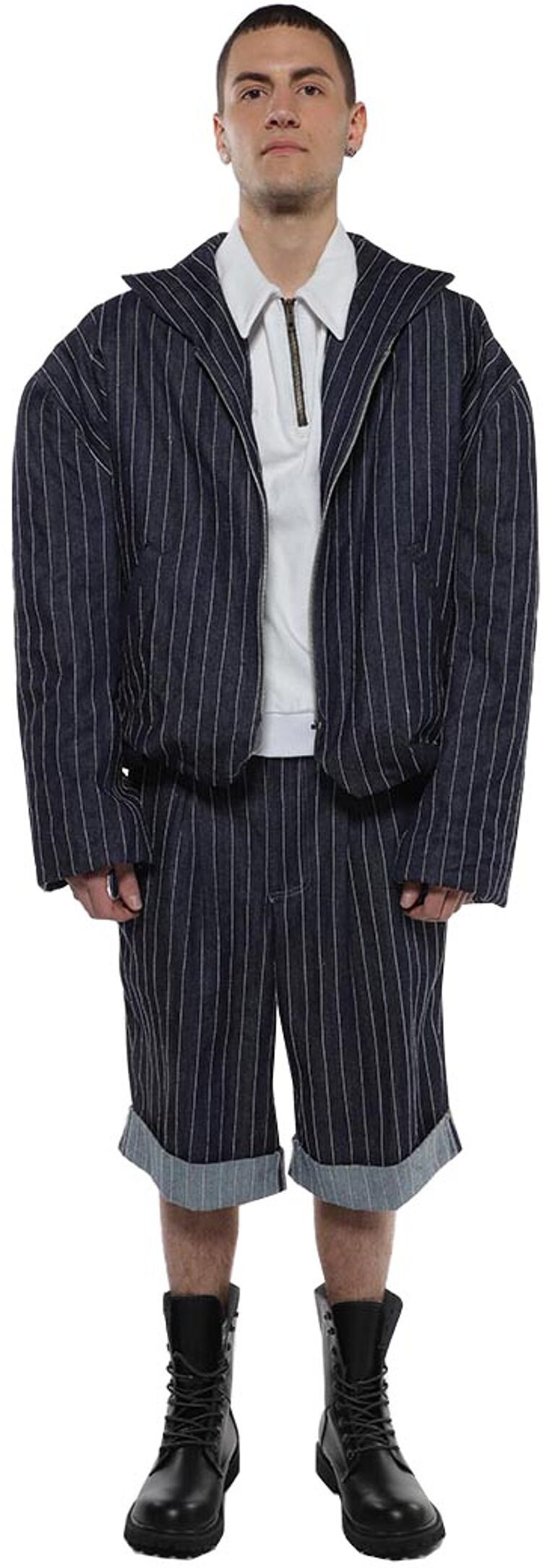 A full shot of a man wearing a pinstripe denim jacket and shorts, paired with a white collared shirt and black combat boots.