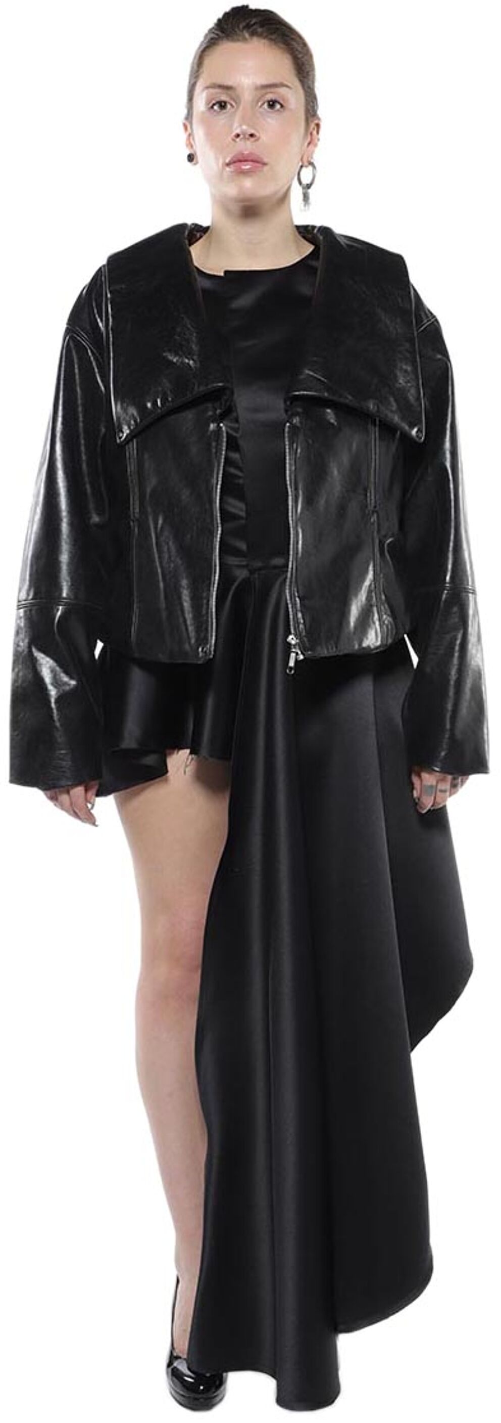 A woman with shoulder-length brown hair wears a black leather jacket over a black asymmetrical satin skirt and black high heels.