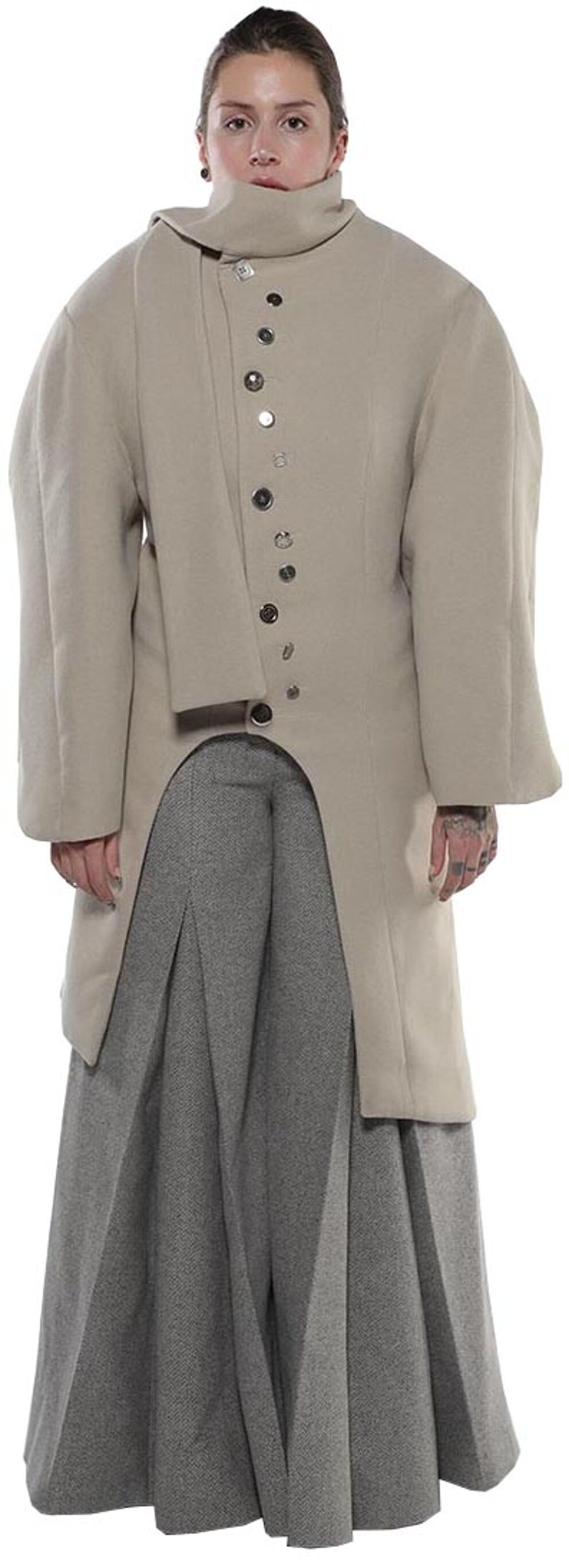 A woman with light skin and dark hair models a long, light beige coat with a high collar and a gray pleated skirt underneath.