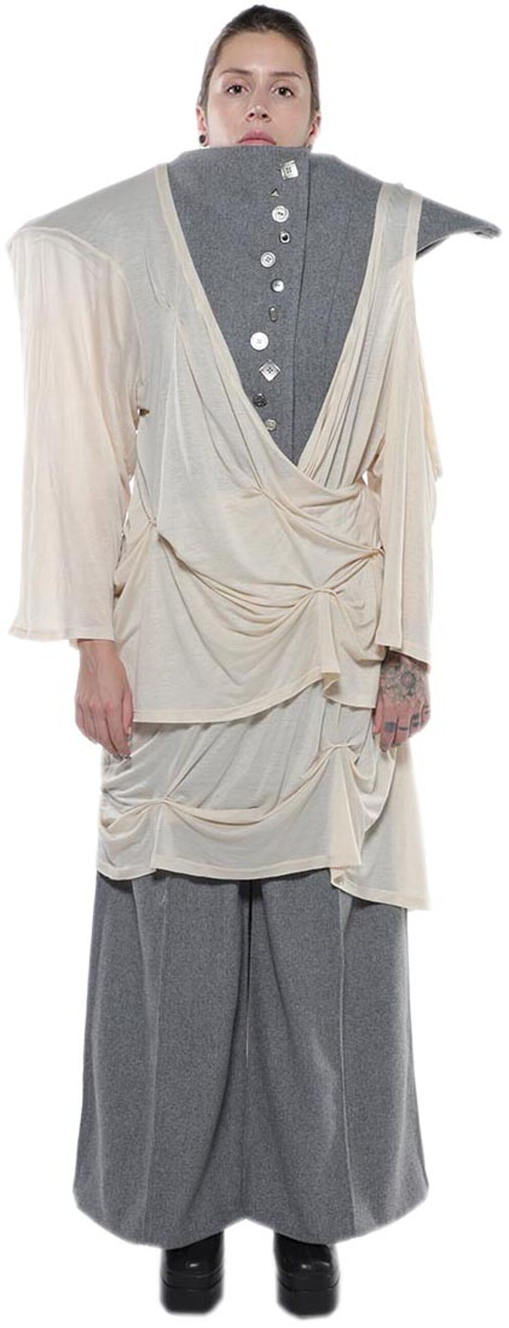 A person with fair skin and dark hair wears a long-sleeved, light beige, draped top over a gray, high-waisted skirt.