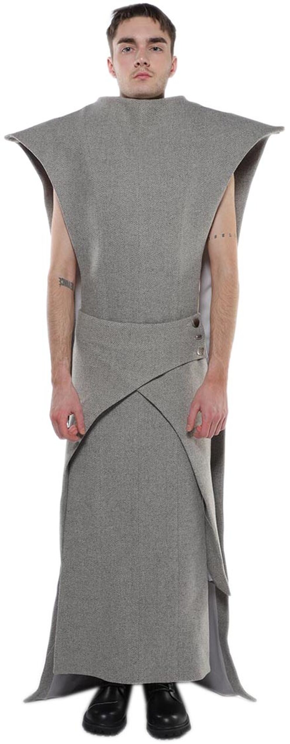 A slender man with short brown hair stands against a white background wearing a long, gray, asymmetrical tunic with a high neckline and cape-like shoulders.