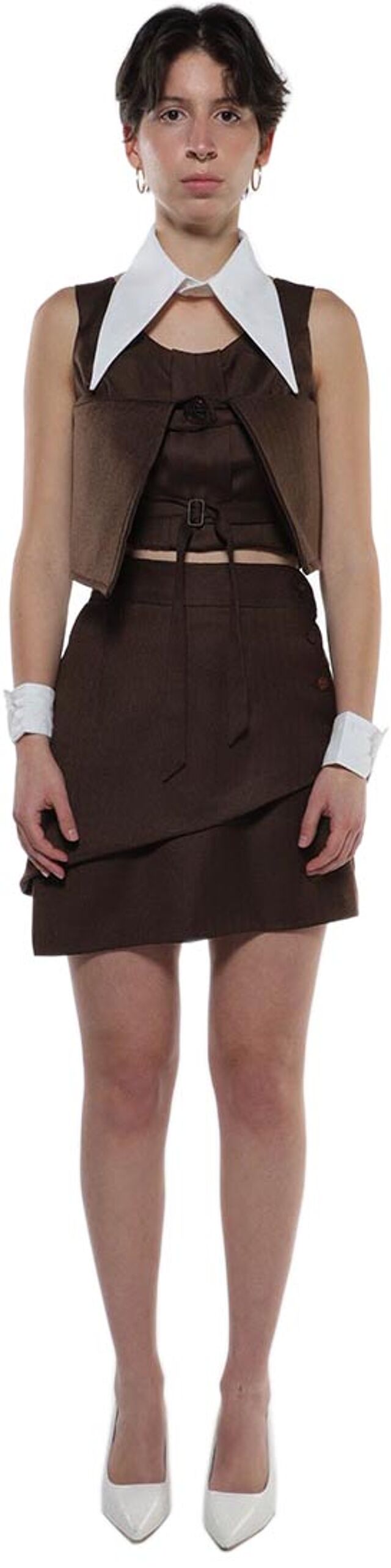 A young woman with short dark hair wears a brown mini skirt, brown vest, and white collared shirt, accessorized with white wristbands and white heels.