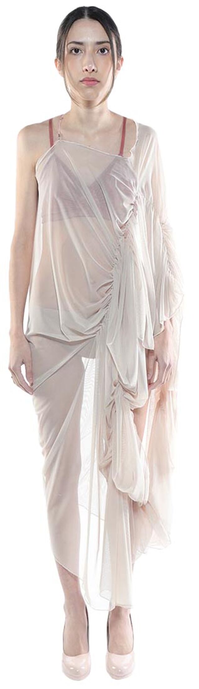 A woman with dark hair pulled back wears a sheer, light beige, asymmetrical draped dress and light pink heels.