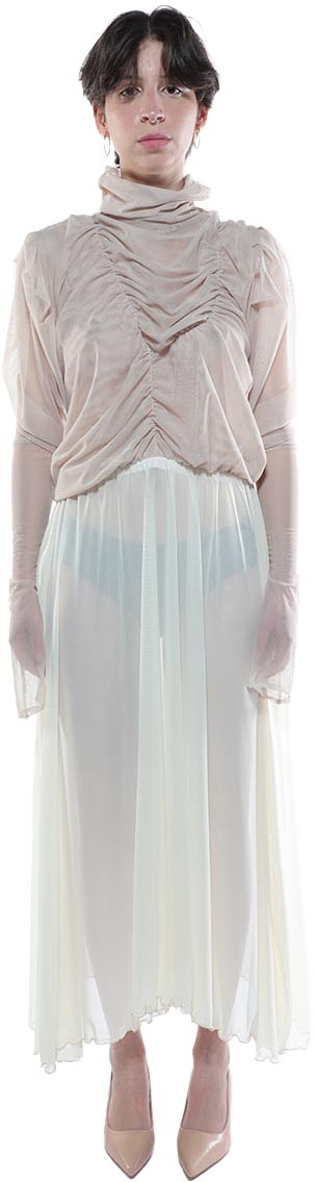 A woman with short dark hair is shown wearing a long, light beige sheer dress with a high neck and long sleeves, and light beige high heels.