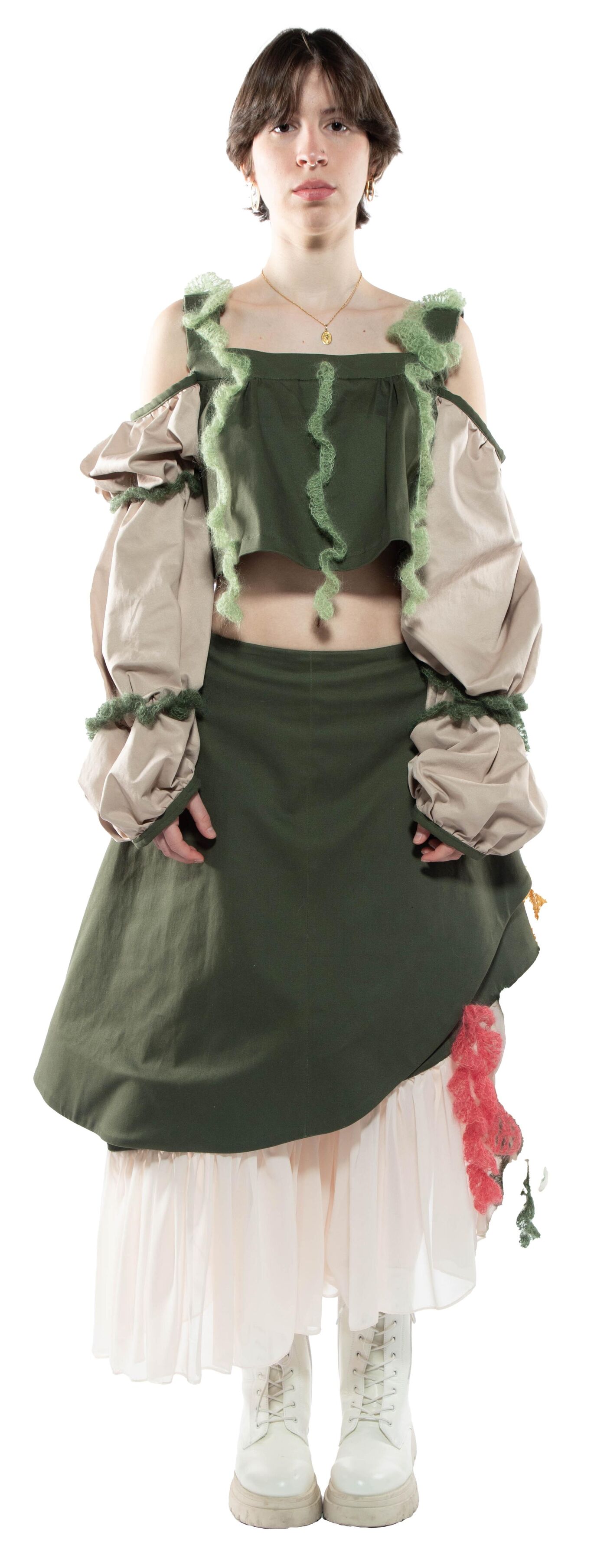 A young woman with short brown hair wears a layered olive green and beige outfit with a ruffled white petticoat and chunky boots.