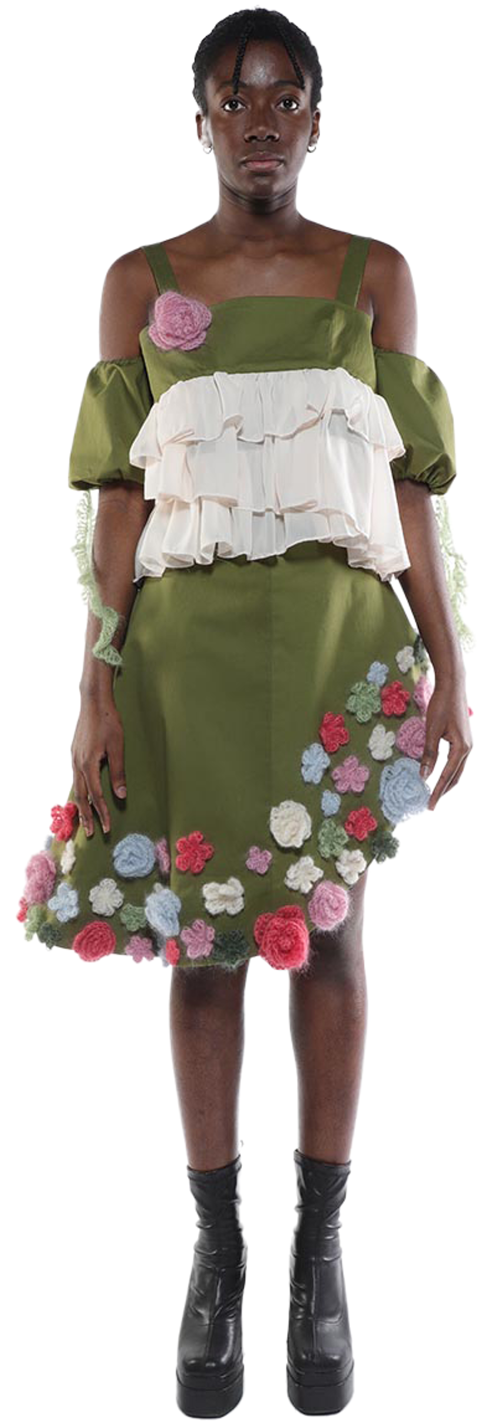 A young Black woman with short dark hair wears a short olive green skirt adorned with crocheted flowers, a layered off-the-shoulder top, and black platform boots.