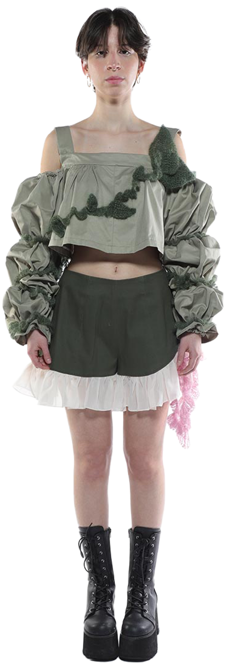 A person with short brown hair wears a sage green off-the-shoulder top with voluminous sleeves, olive green shorts with a white ruffle, and black platform boots.