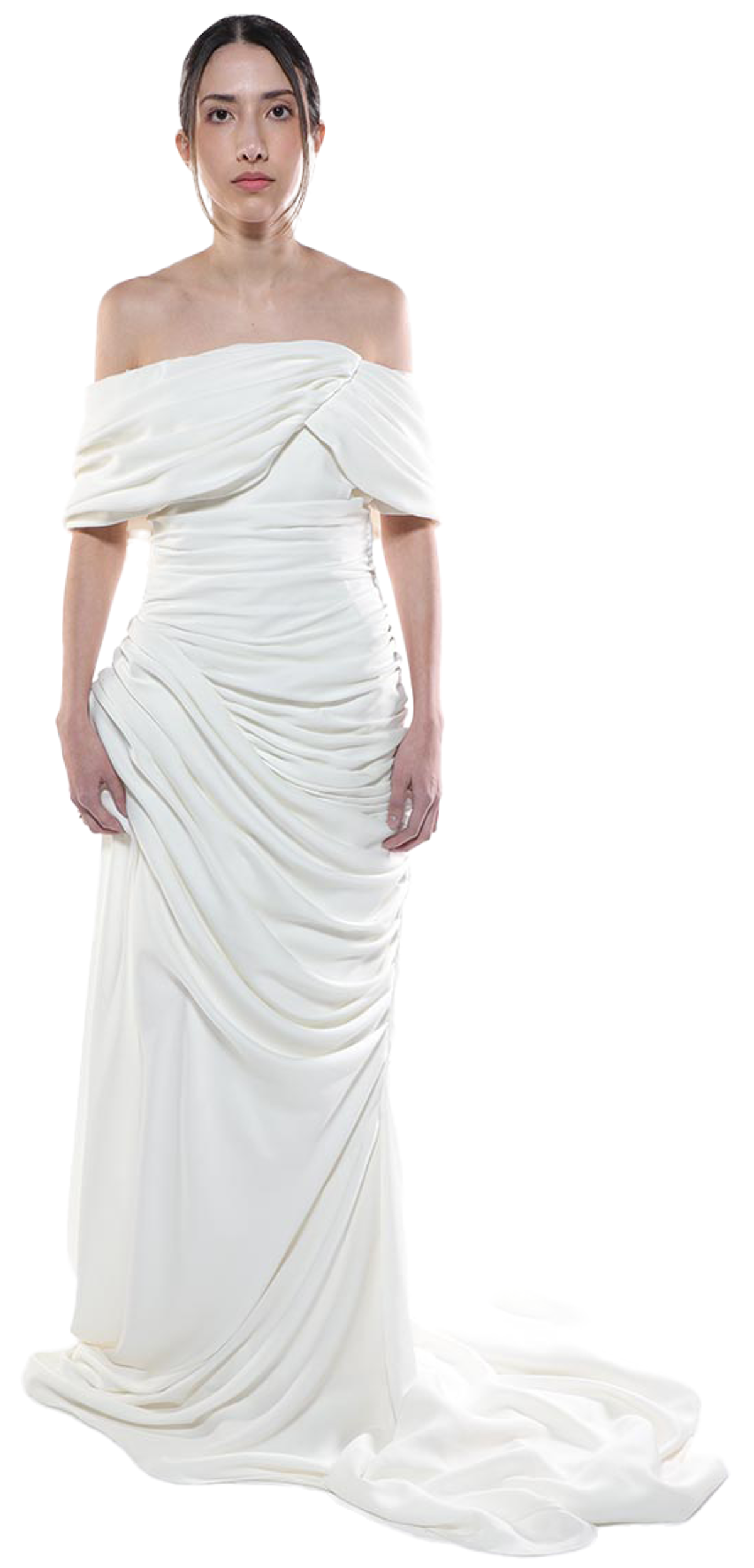A woman with dark hair pulled back stands against a white background, wearing an off-the-shoulder, floor-length white gown. The bodice is fitted, with draped fabric that continues to the hem, creating a flowing, elegant look. The fabric appears smooth and possibly satin-like. Her expression is serious. The dress has a train that pools on the floor.