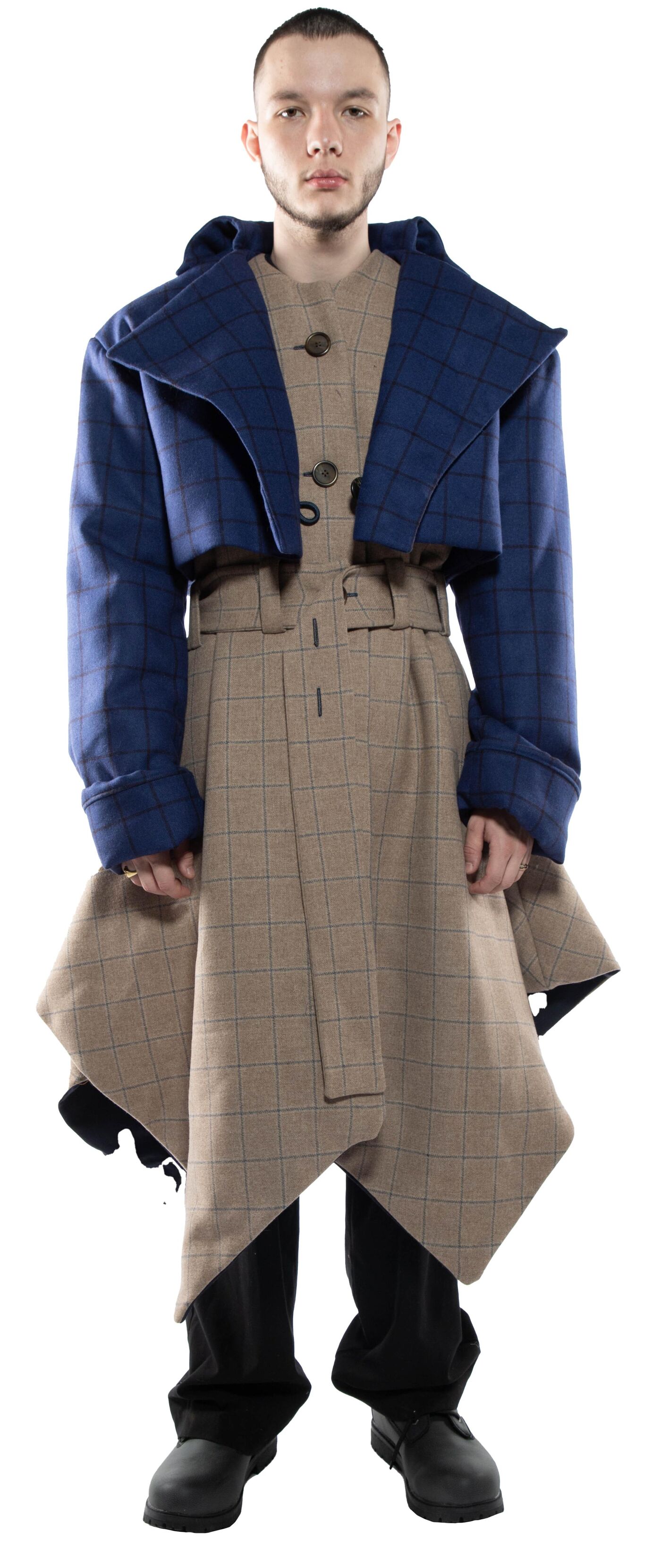 A person wearing a blue cropped jacket over a beige plaid asymmetrical skirt and black pants stands against a white background.