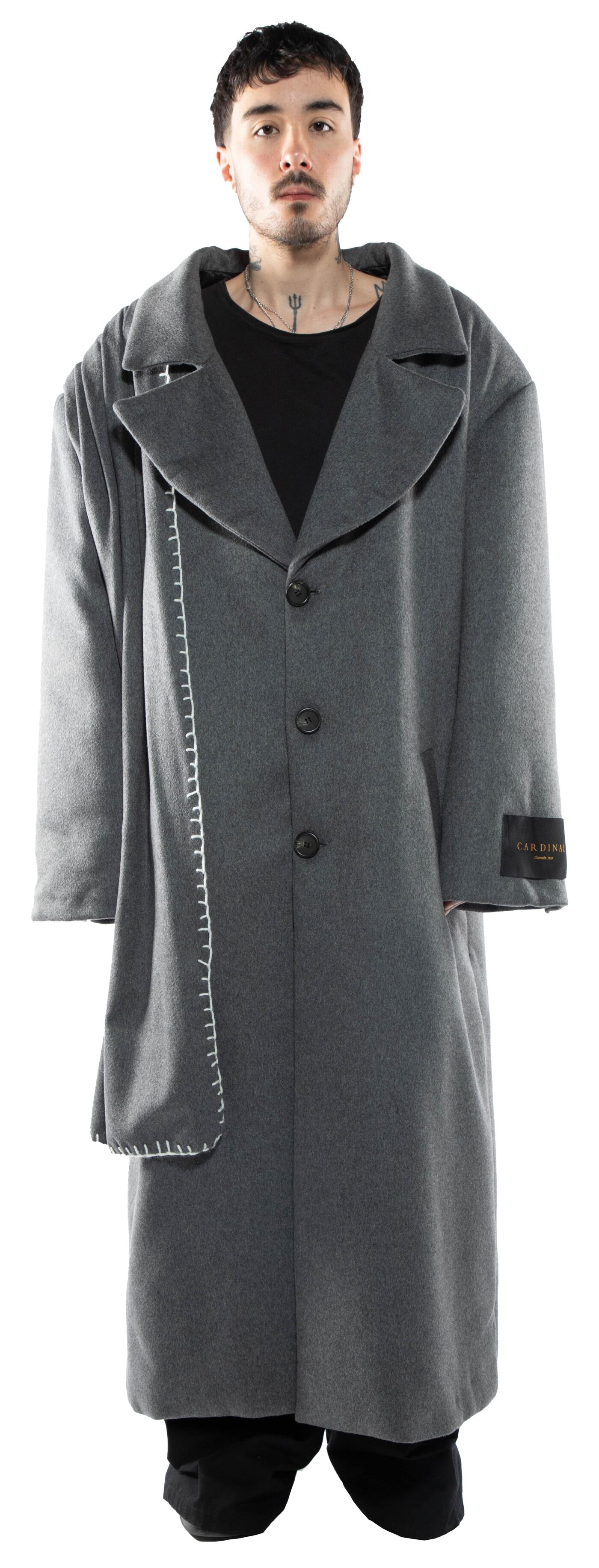 A man with dark hair and tattoos stands against a white background wearing a long, gray, oversized coat with a contrasting lighter gray fabric detail on the left side.