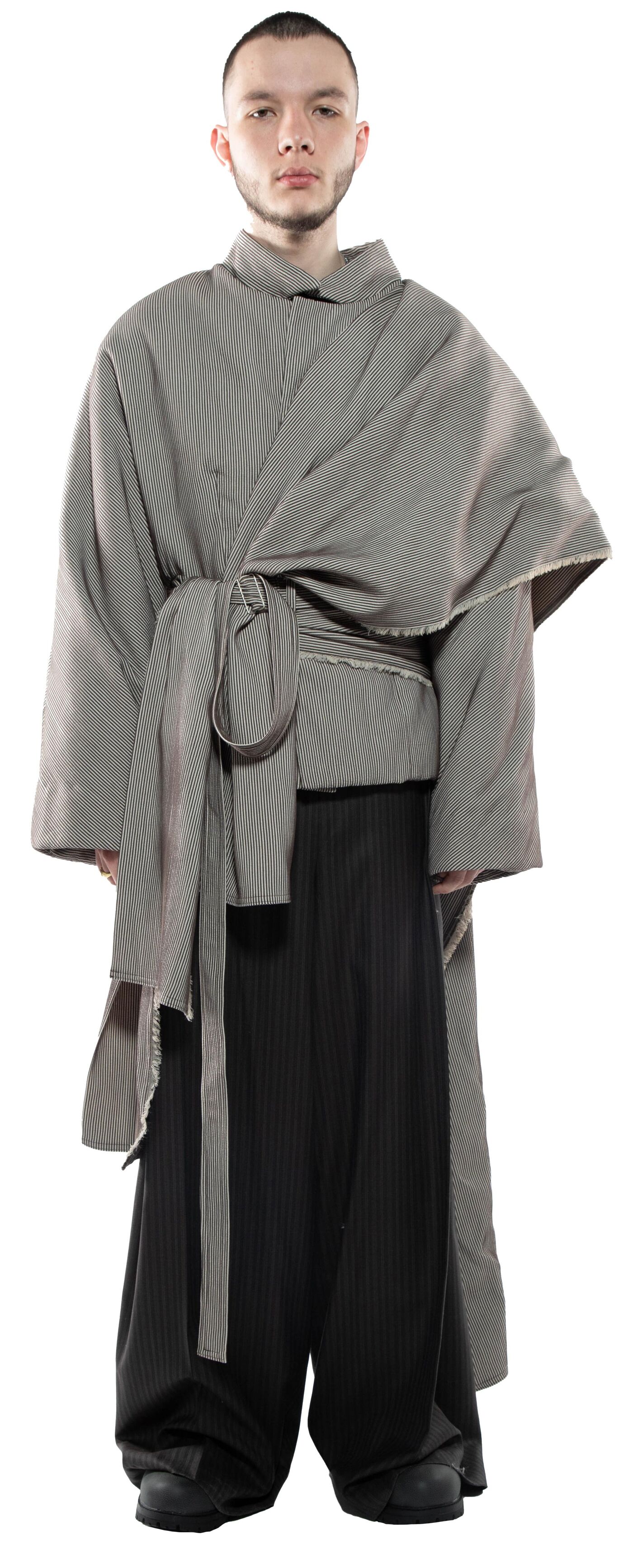 A person with short brown hair wears a long, light-gray, draped coat over dark-gray, wide-legged pants.