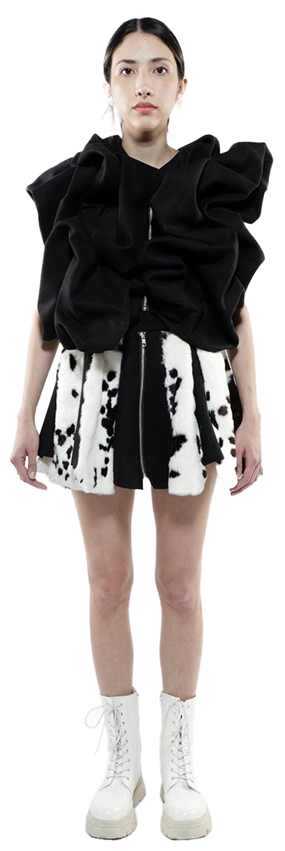 A young woman with dark hair pulled back in a ponytail stands against a white background wearing a black, ruffled cropped top, a black and white patterned mini-skirt, and white lace-up combat boots.