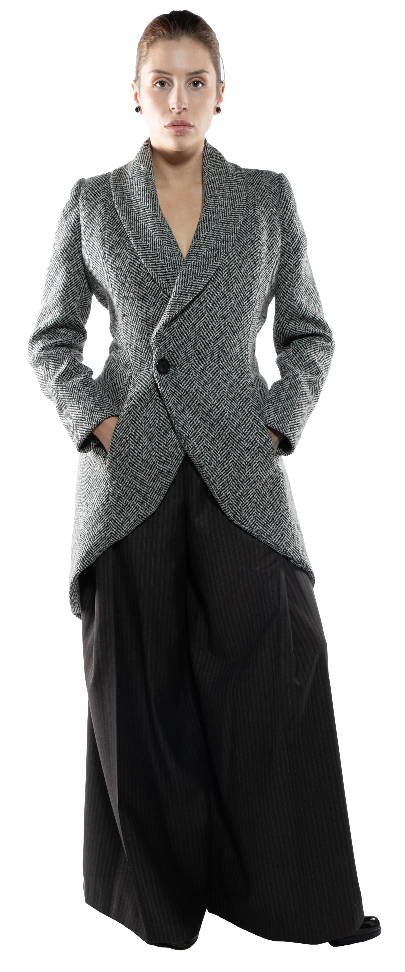 A woman with dark hair pulled back in a bun wears a gray tweed jacket with a long, asymmetrical tail and black pinstriped wide-leg trousers.