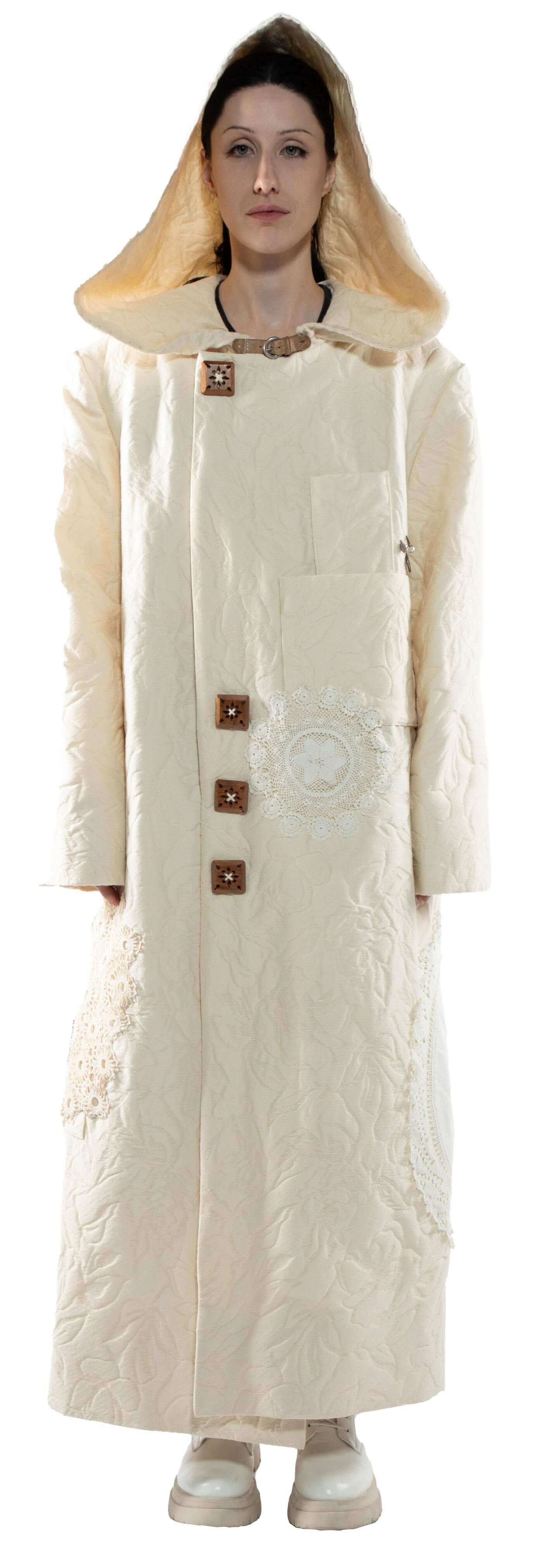 A woman stands wearing a long, off-white coat with a large hood, wooden buttons, and lace detailing.