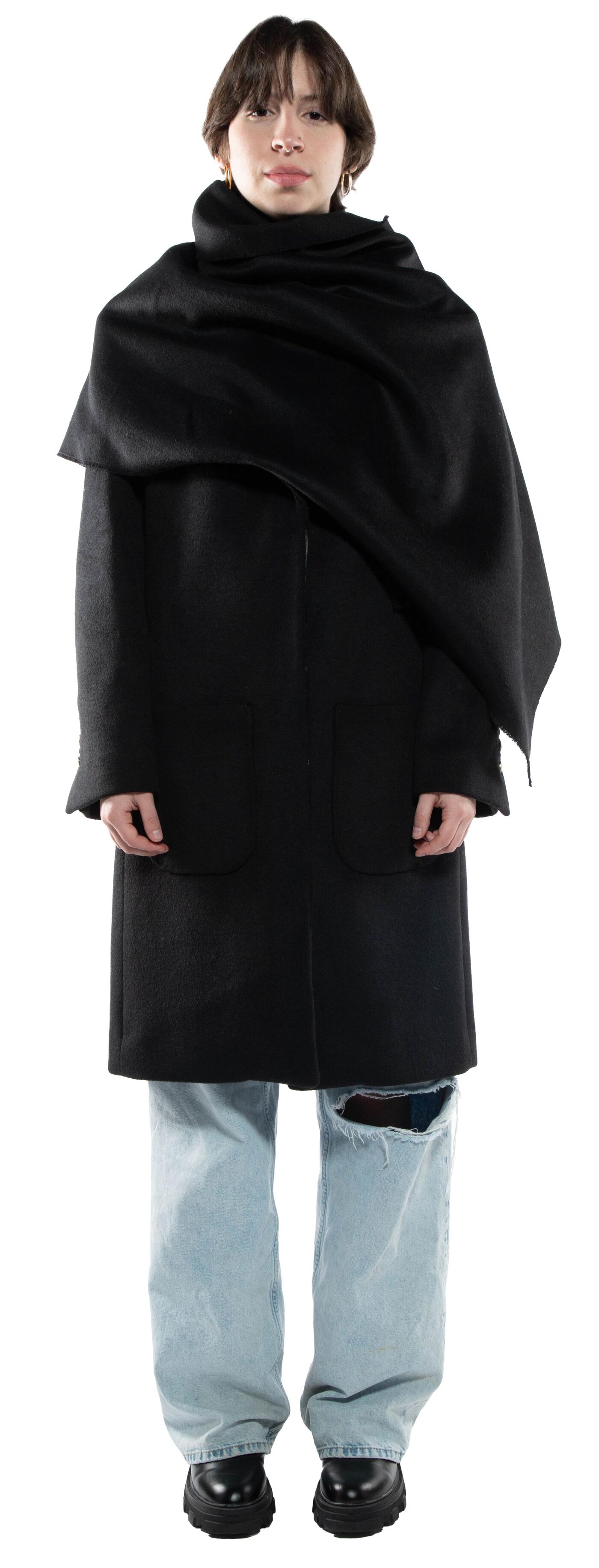 A person with short brown hair wears a long black coat and a large black scarf over it, paired with light blue jeans and black shoes.