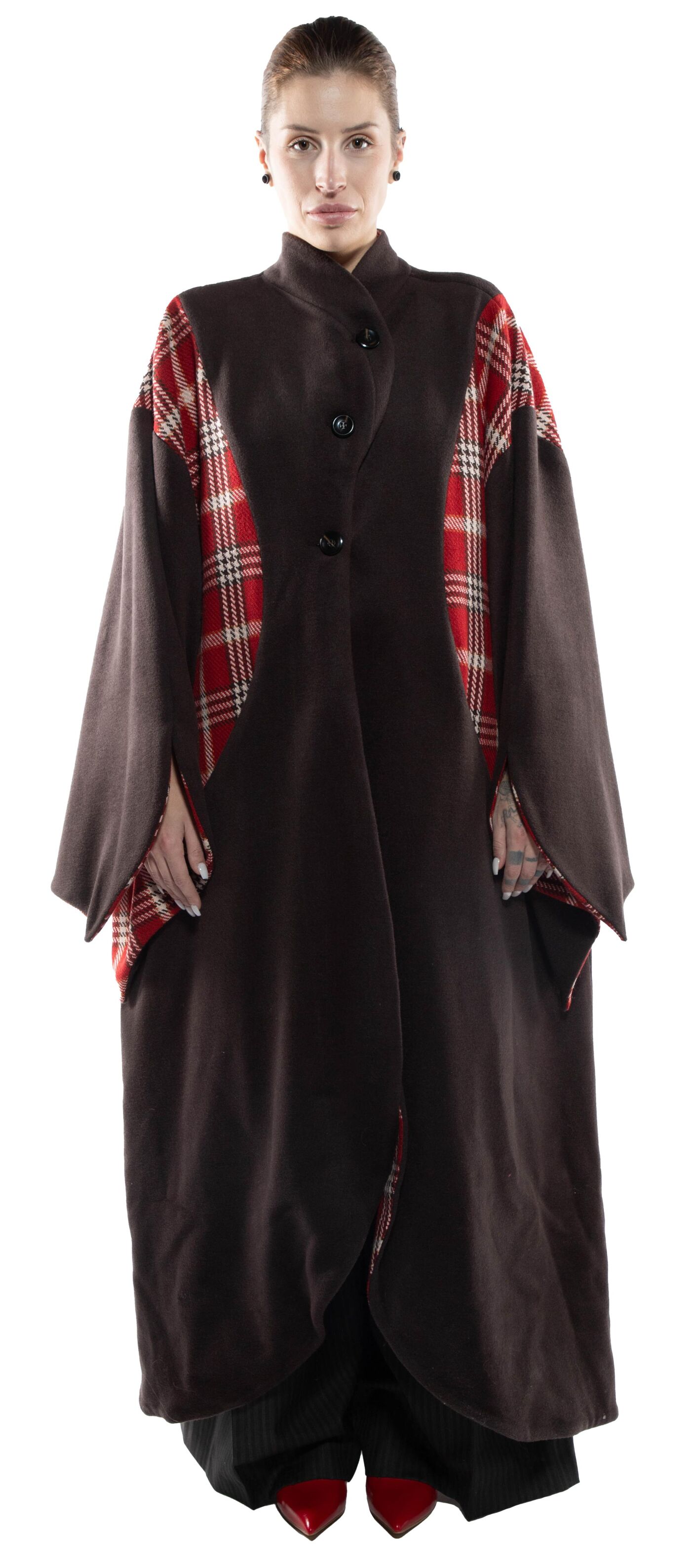 A woman with dark hair pulled back wears a long, dark brown coat with red and white plaid accents at the shoulders and sleeves, paired with dark pants and red shoes.