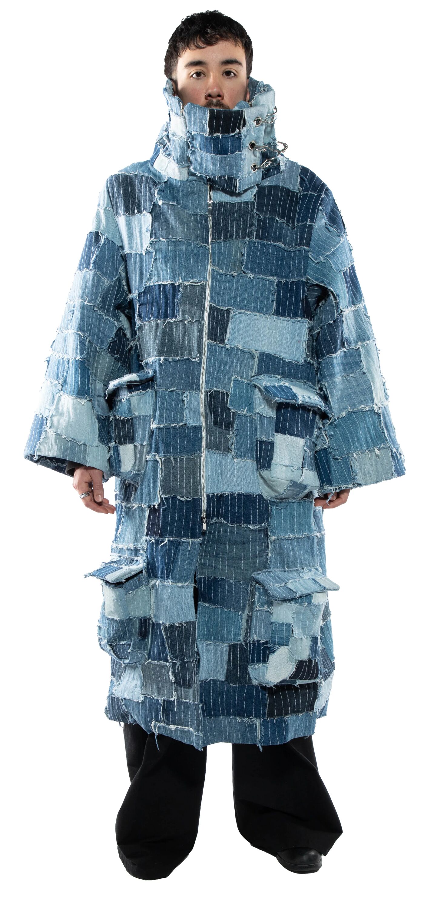 A person with short dark hair stands against a white background wearing a long, patchwork denim coat with a high collar and black pants.