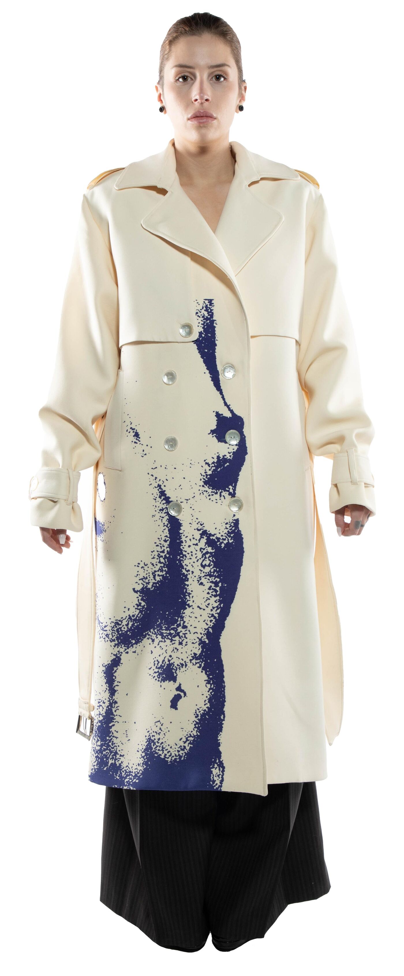 A woman with light skin and dark hair models a long, off-white trench coat with a navy blue abstract print down the front, paired with dark-colored pants.