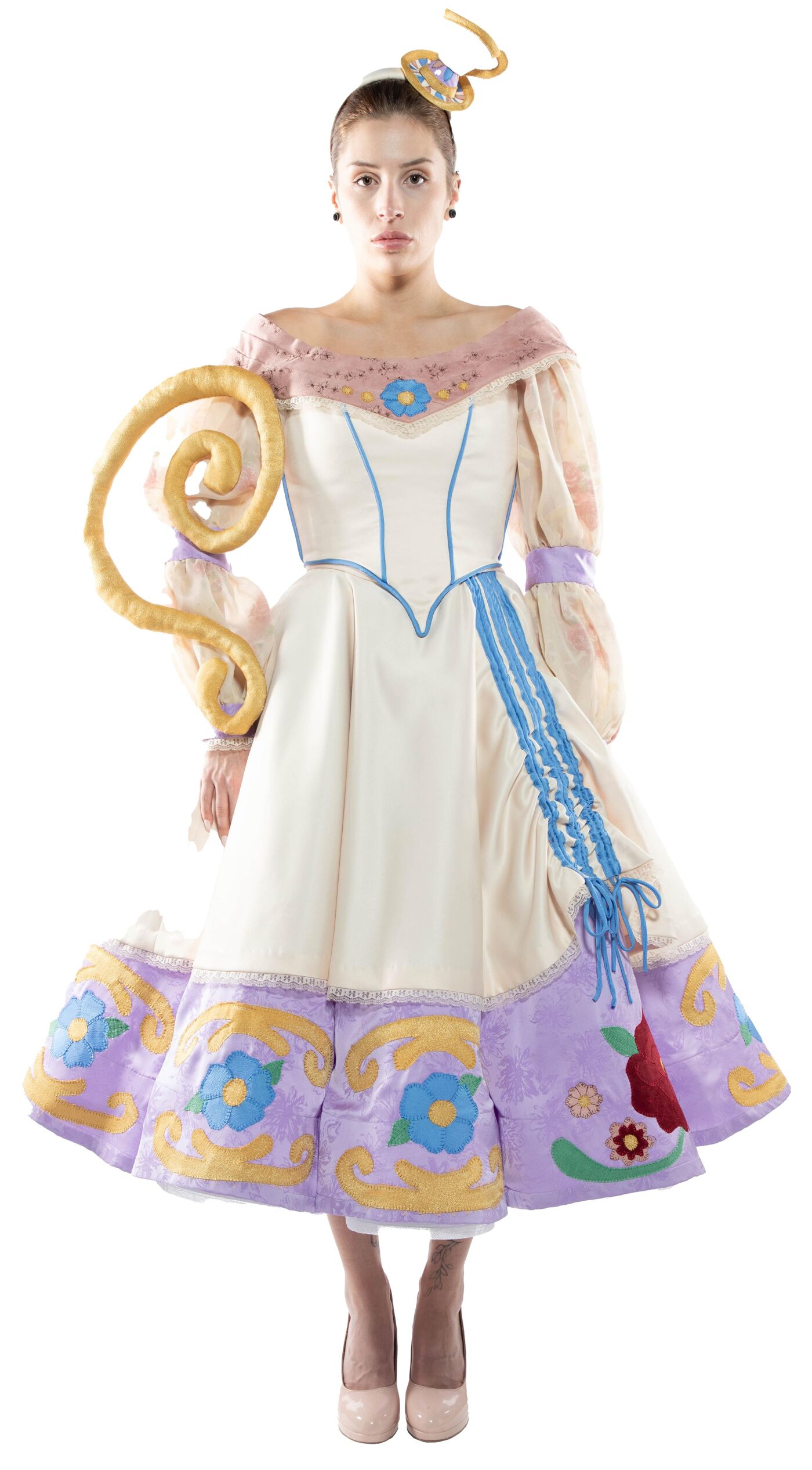 A woman in a white and lavender dress with floral embroidery and a curled pastry-like accessory stands against a white background.