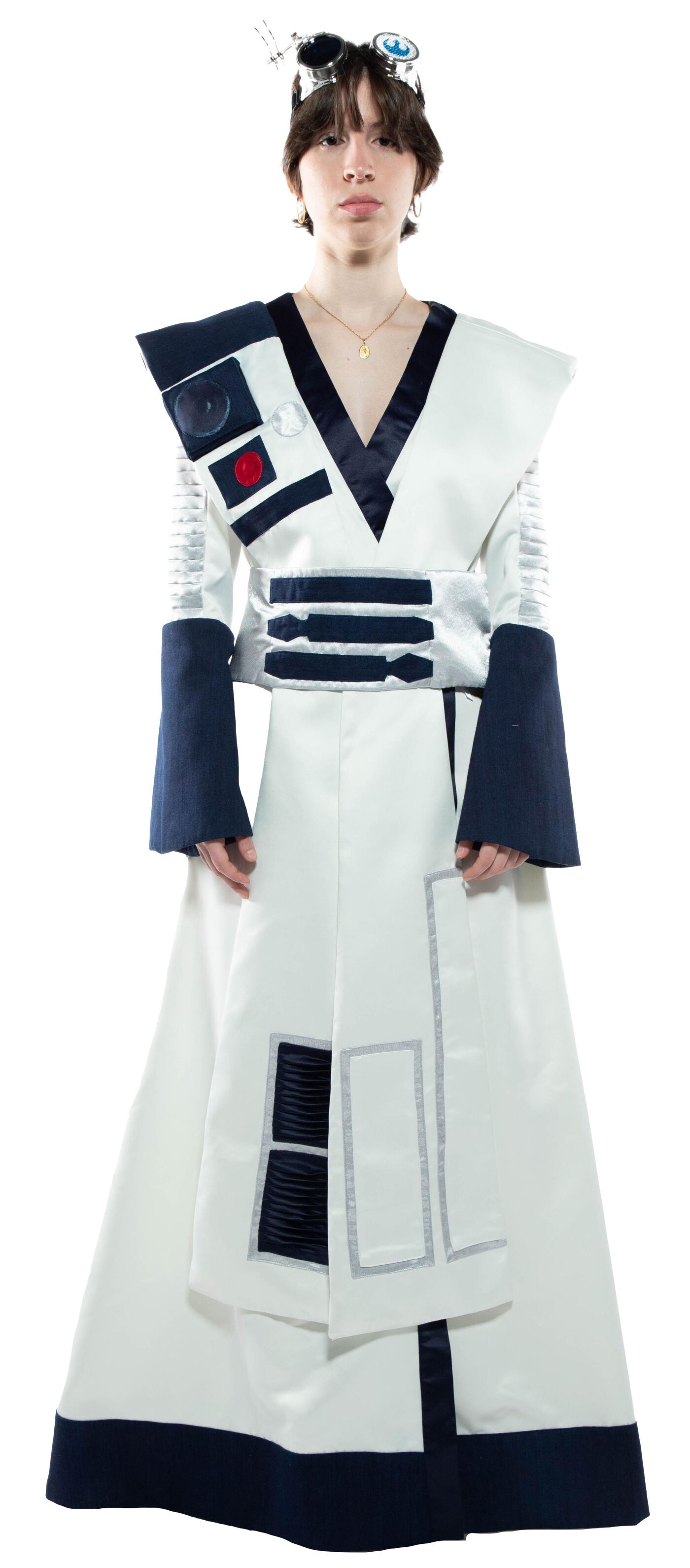 A person in a long, white robe with dark blue accents and a headpiece stands against a white background.