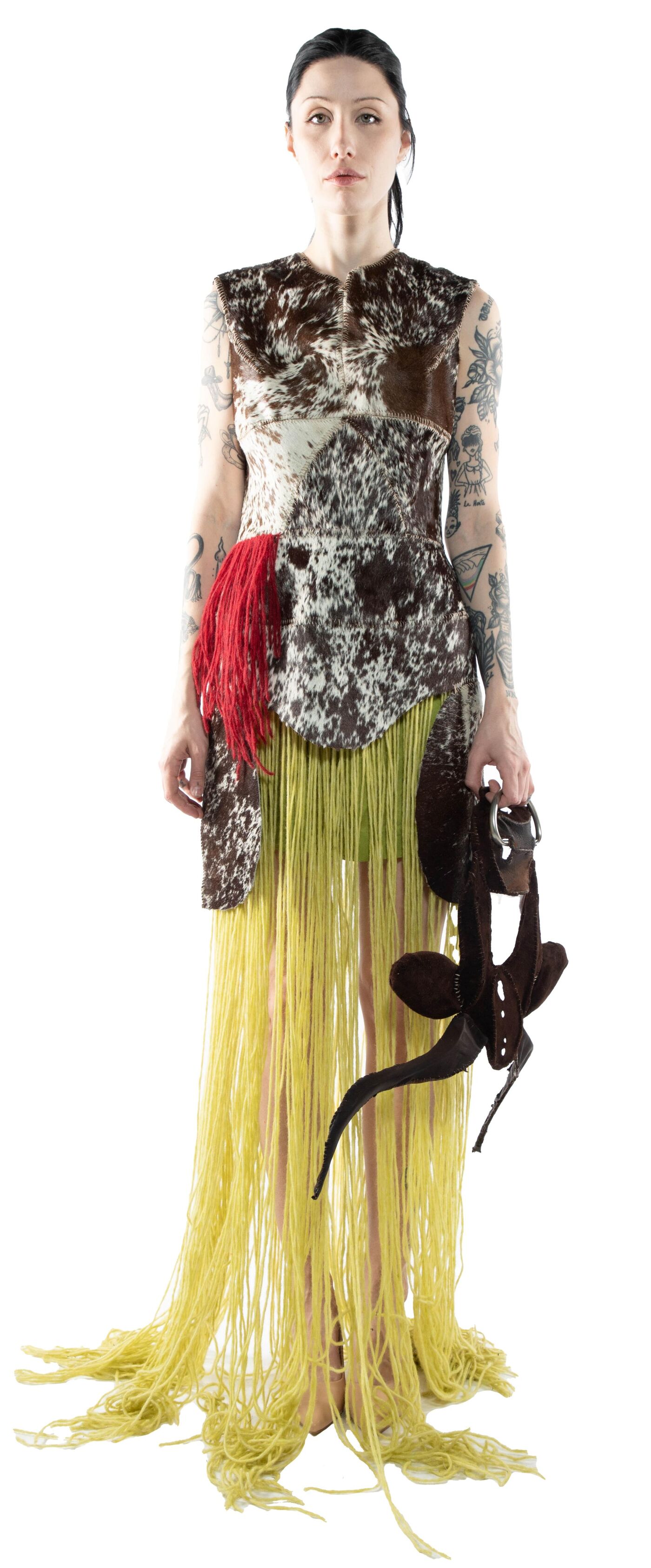 A woman with tattoos stands against a white background, wearing a calf-length, fringed dress with a cowhide pattern, holding a pair of dark brown shoes.