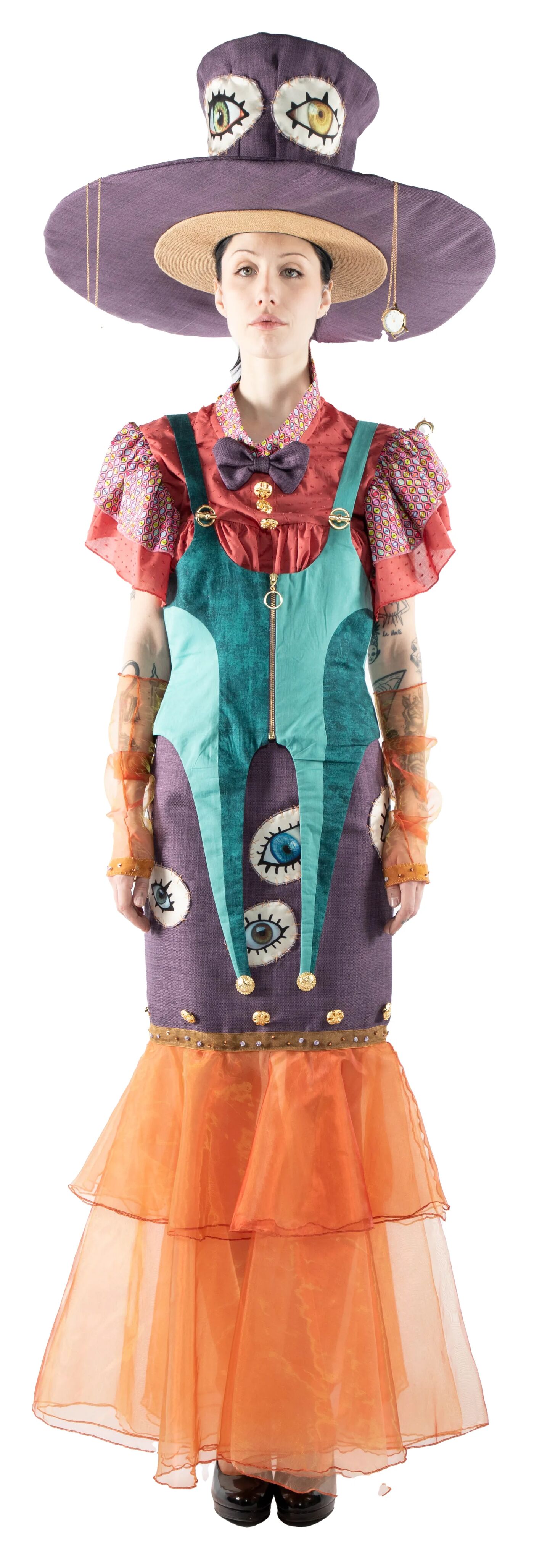 A full shot of a model wearing a whimsical outfit consisting of a large, eye-decorated hat, a multicolored dress with a teal bodice and orange tulle skirt, and orange sheer gloves.