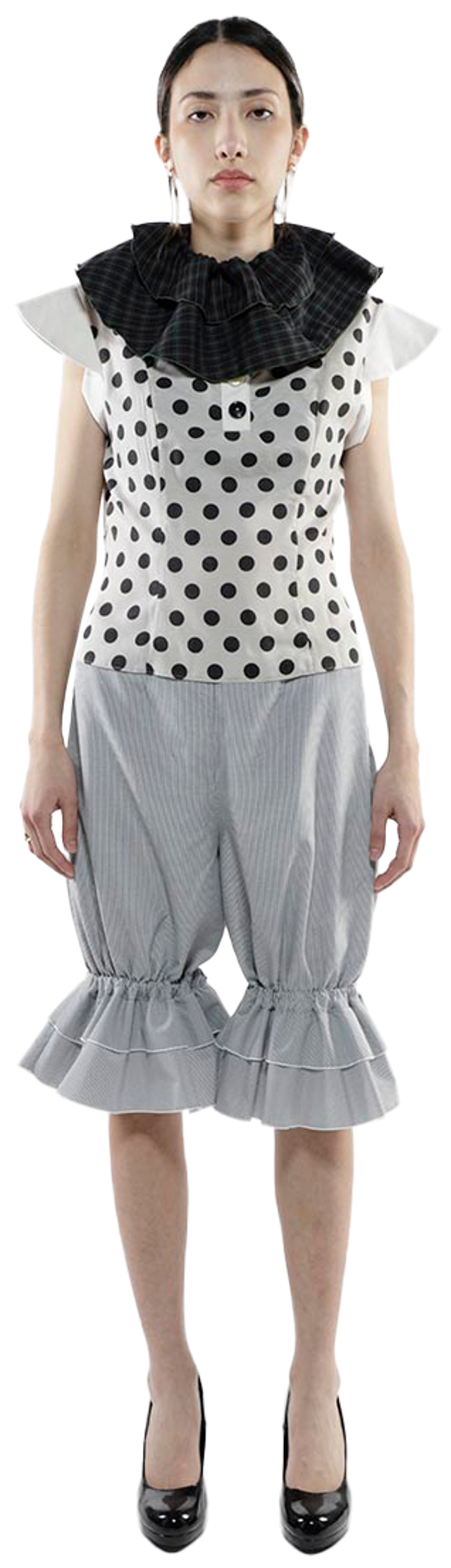 A full shot of a woman in a white polka dot vest, ruffled grey bloomers, a black ruffled collar, and black heels, standing against a white background.