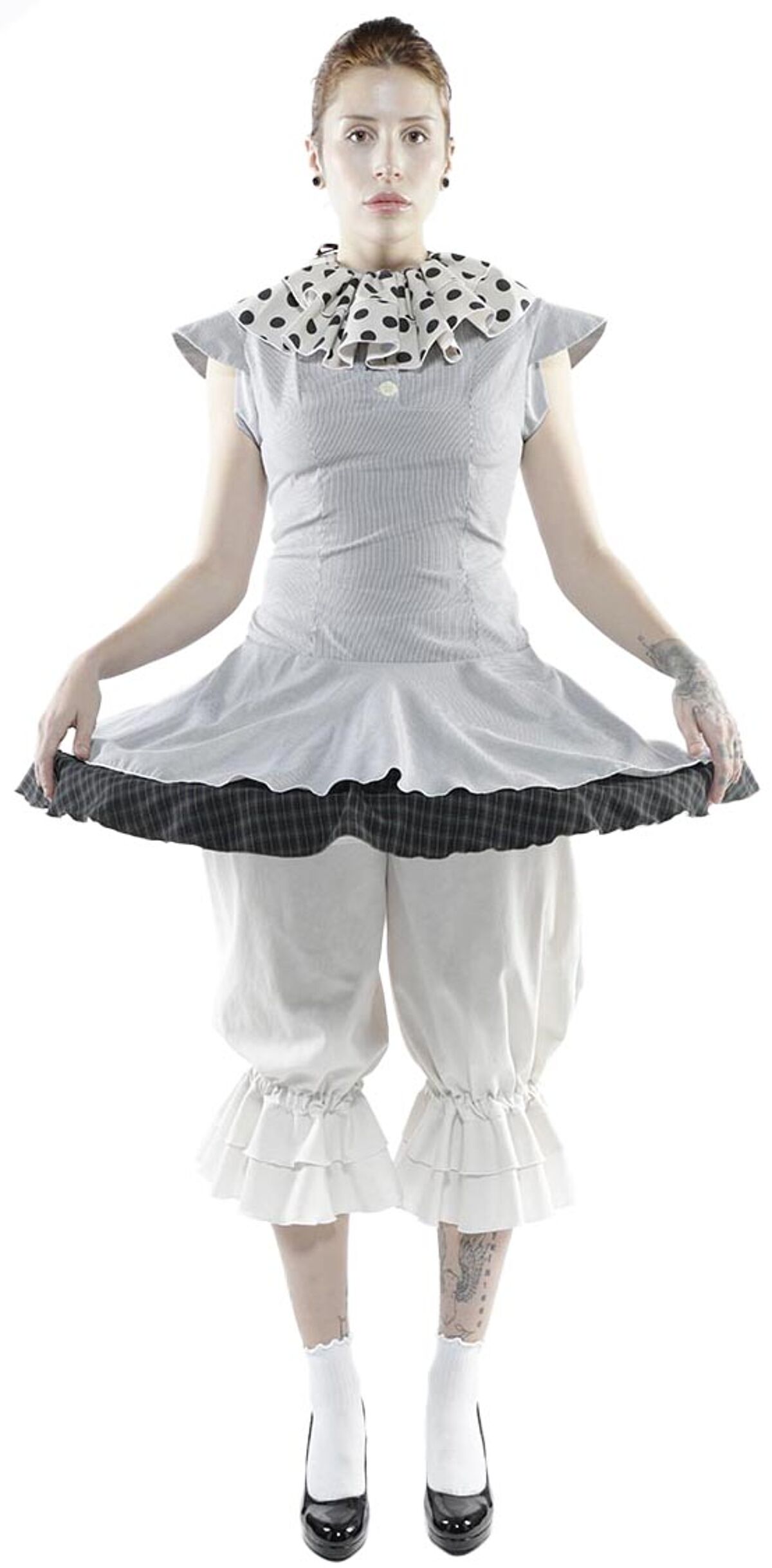 A fair-skinned woman with a pale face and dark hair, wearing a grey and white polka dot dress with white bloomers and black shoes, stands against a white background.