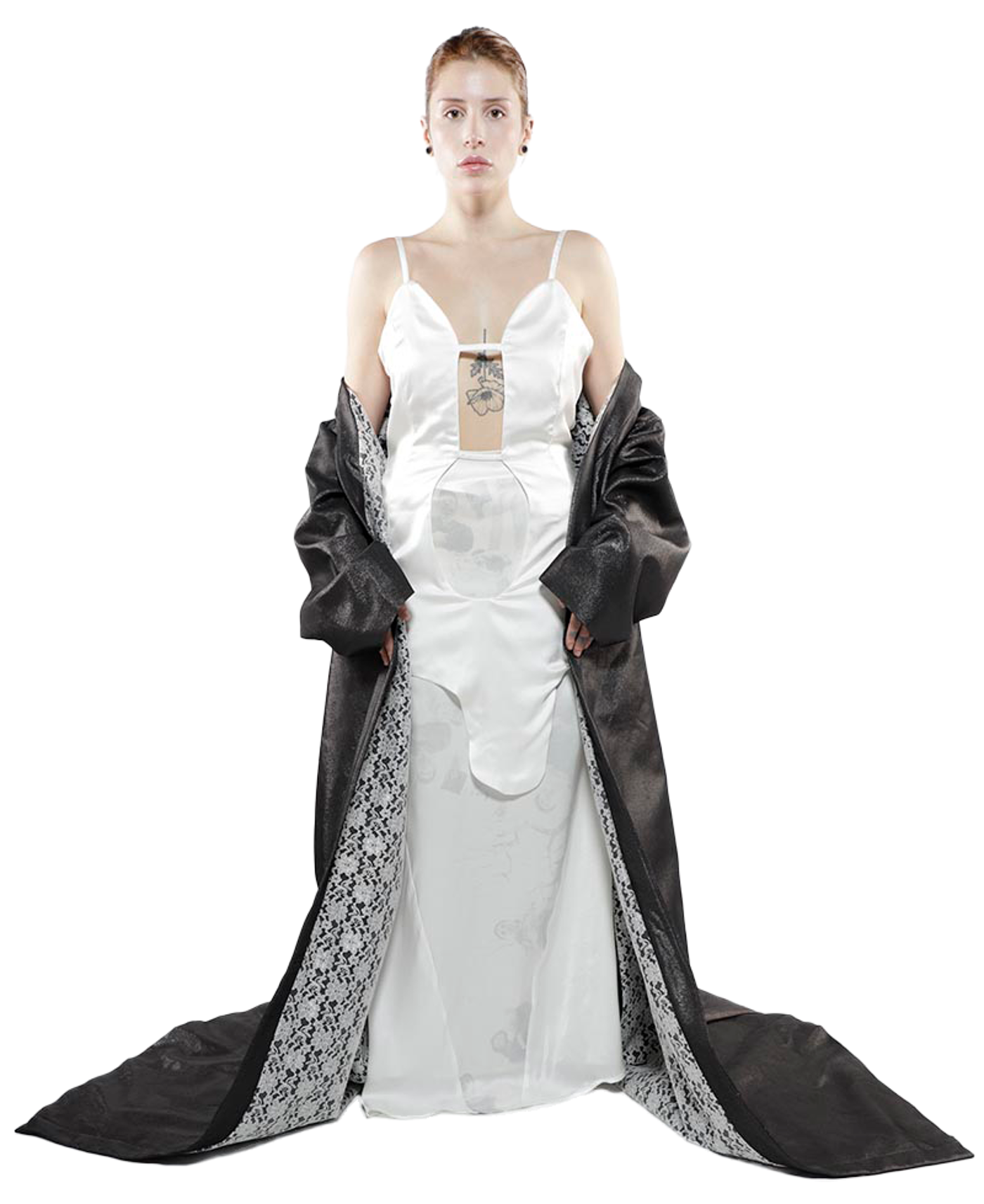A woman with short, reddish-brown hair stands against a white background, wearing a white satin dress with cutouts and a long, dark gray coat with lace detailing, posing directly at the camera.